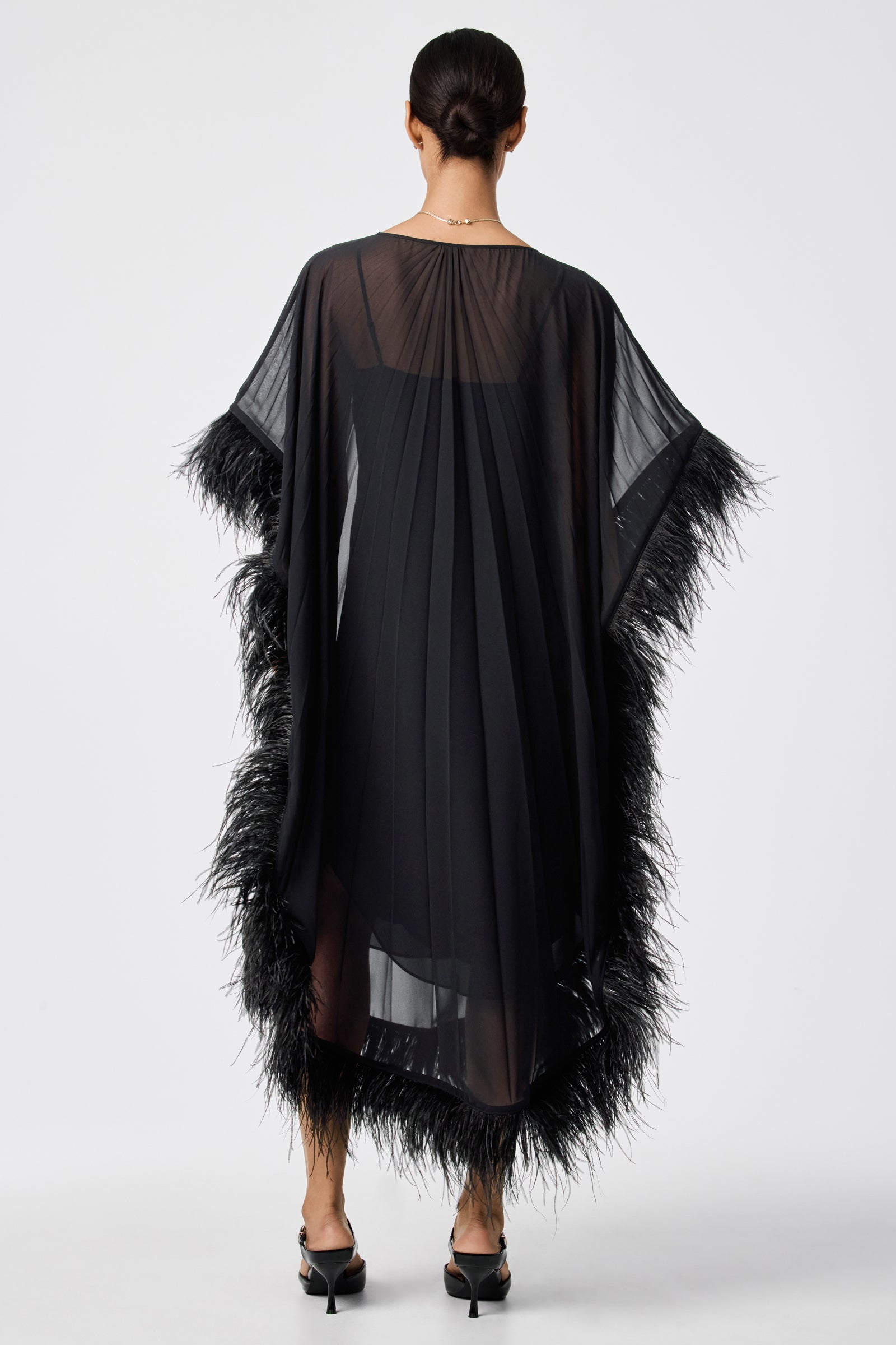 A woman with her hair in a low bun faces away, wearing the sheer black Pleated Neena Feather-Trimmed Caftan Dress with feather trim on the sleeves and hem, paired with black heeled sandals against a plain white background.
