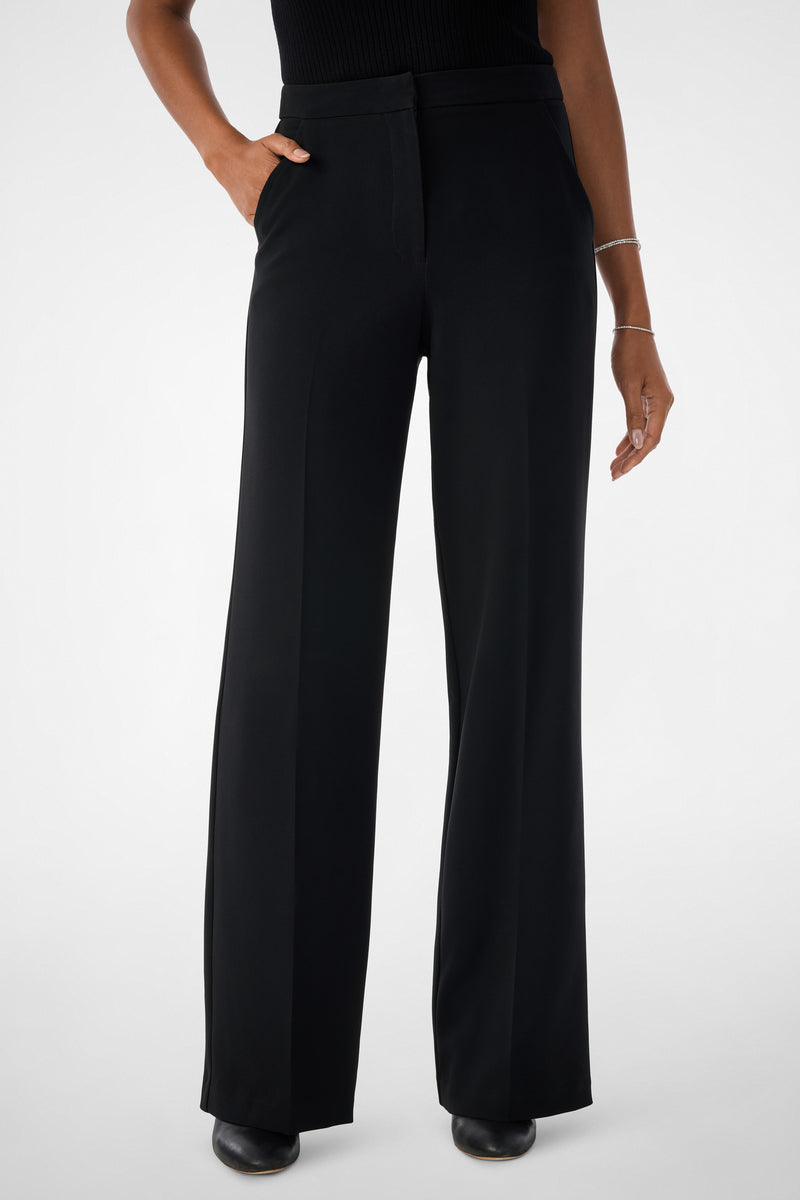 Wearing the Sadie Wide-Leg Pants in black paired with a black sleeveless top and black shoes, one hand is in a pocket while the other arm rests by their side.