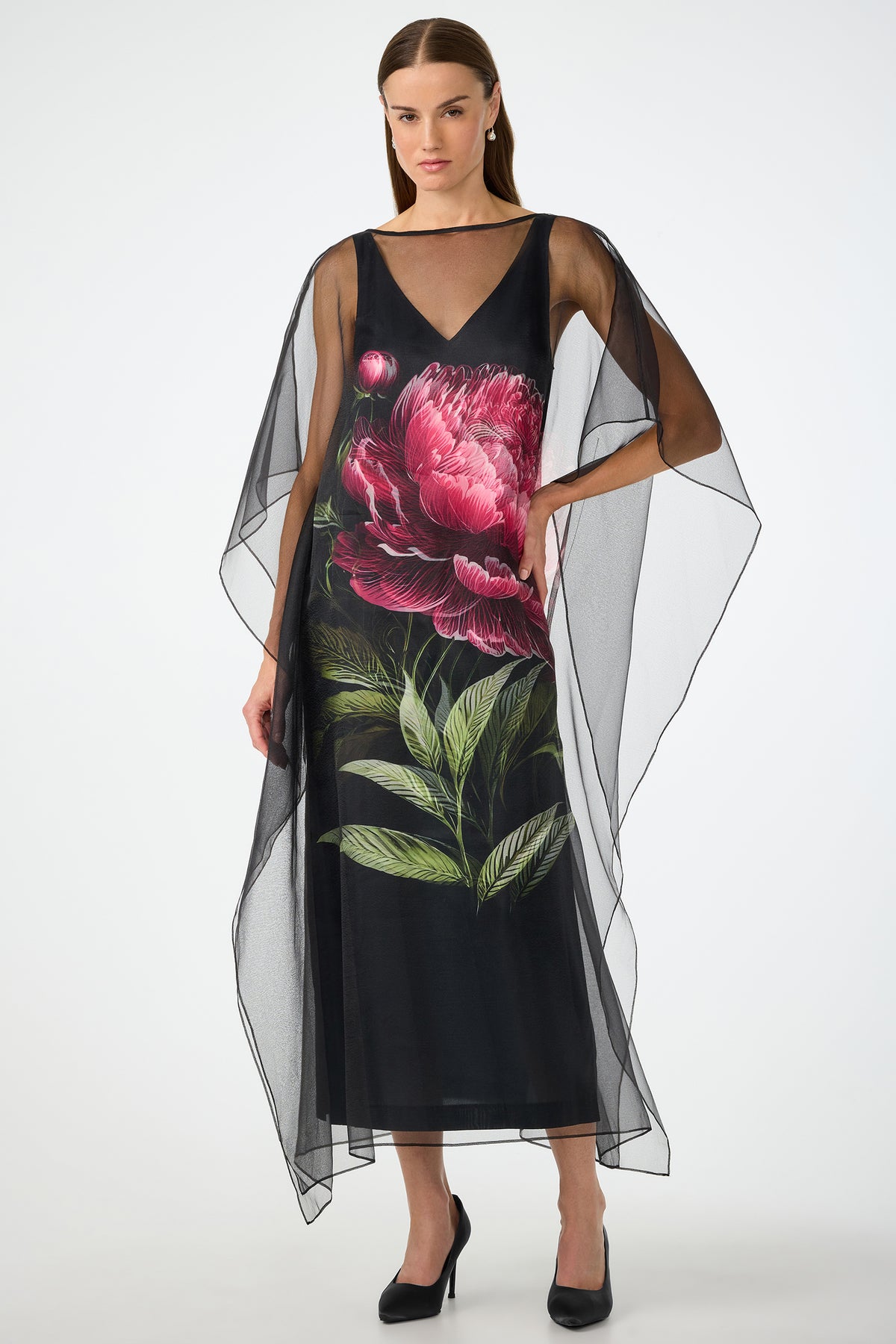 A woman wears the Salon 2 Organza Midi Dress in Fauna Floral, a sheer black cape dress with wide, flowing sleeves and bold pink peony and green leaf print on the front. She stands confidently in black heels with one hand on her hip.