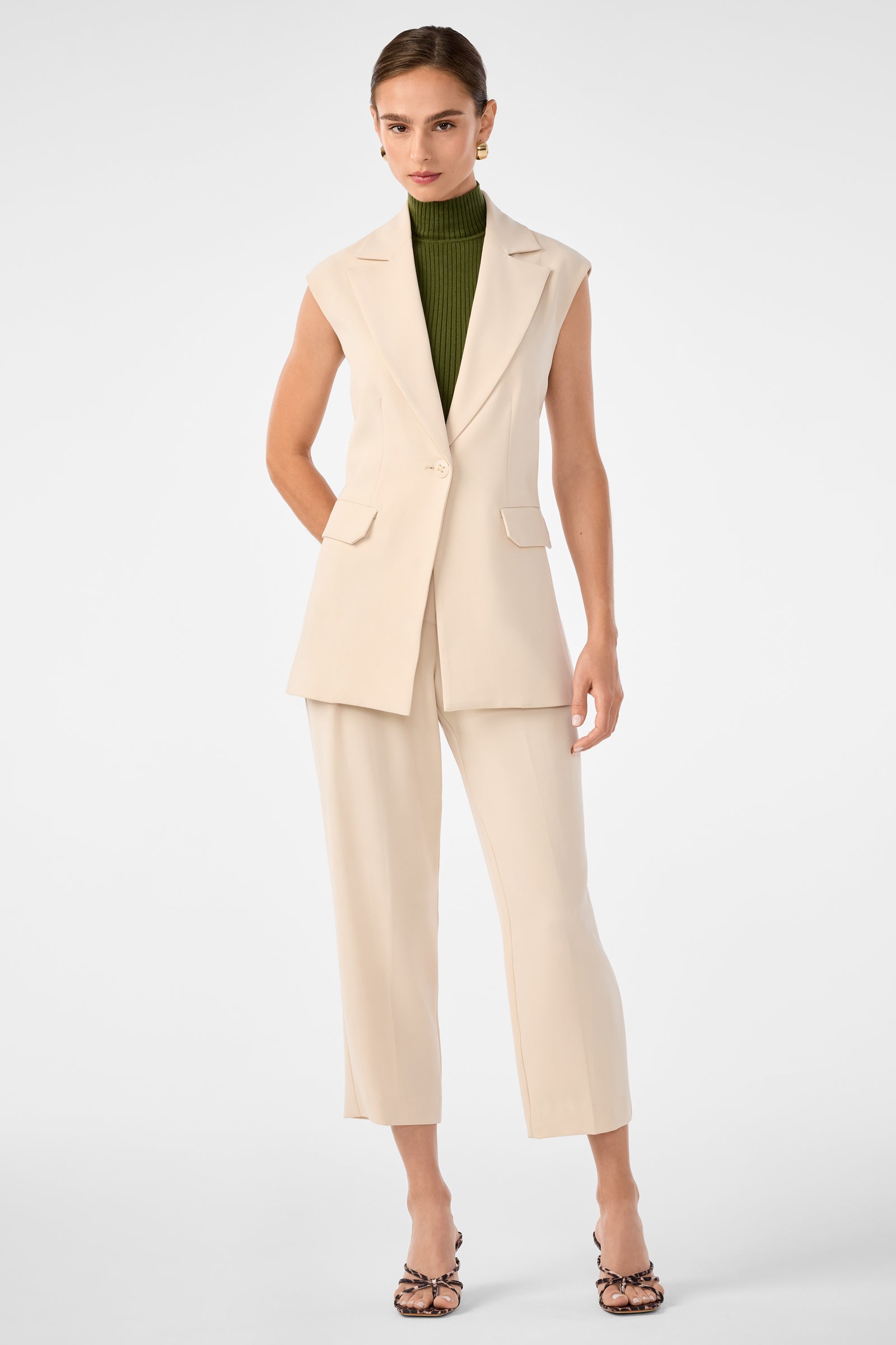 A woman poses against a plain background wearing the Boabab Sleeveless Jacket over a green turtleneck, paired with beige pants and patterned heeled sandals. Her hair is pulled back and she stands with one hand in her pocket.