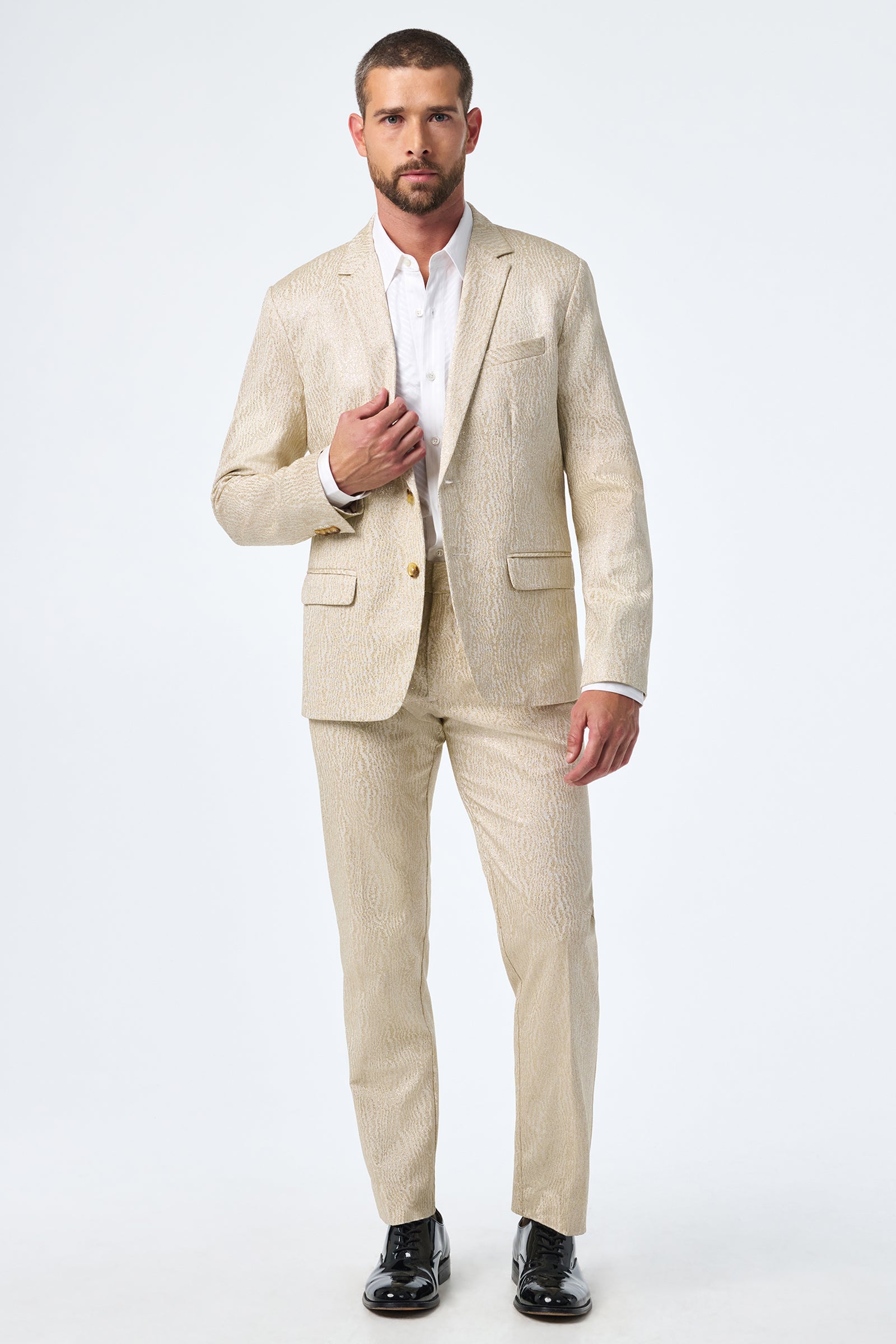 A man with short hair and a beard models the Clyde Slim Trouser in a light beige patterned suit, paired with a white dress shirt and black shoes, standing against a plain white background.