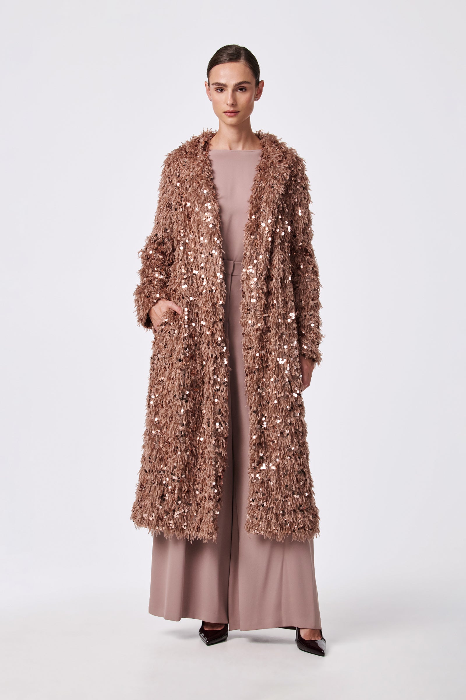 A woman stands against a plain background, wearing the Anna Coat—a long, rose-gold sequin-textured coat—over a blush pink wide-leg jumpsuit and black pointed heels. Her hair is neatly pulled back.