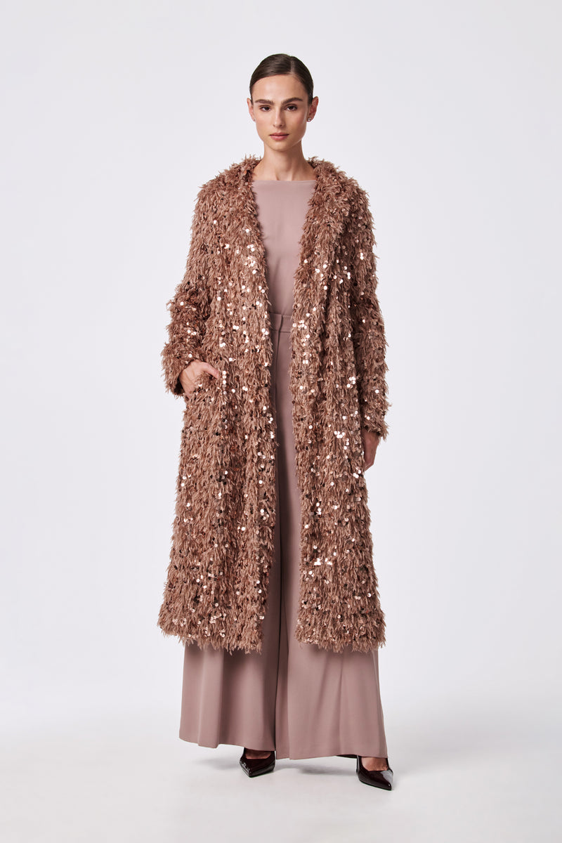 A woman stands against a plain background, wearing the Anna Coat—a long, rose-gold sequin-textured coat—over a blush pink wide-leg jumpsuit and black pointed heels. Her hair is neatly pulled back.