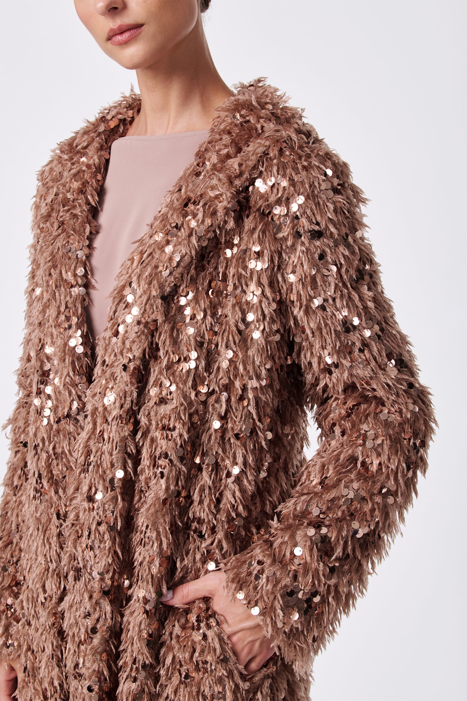 A woman wears the Anna Coat, a textured light brown faux fur piece adorned with shiny sequins. With one hand in her pocket and her face partially visible, she poses against a plain white background.