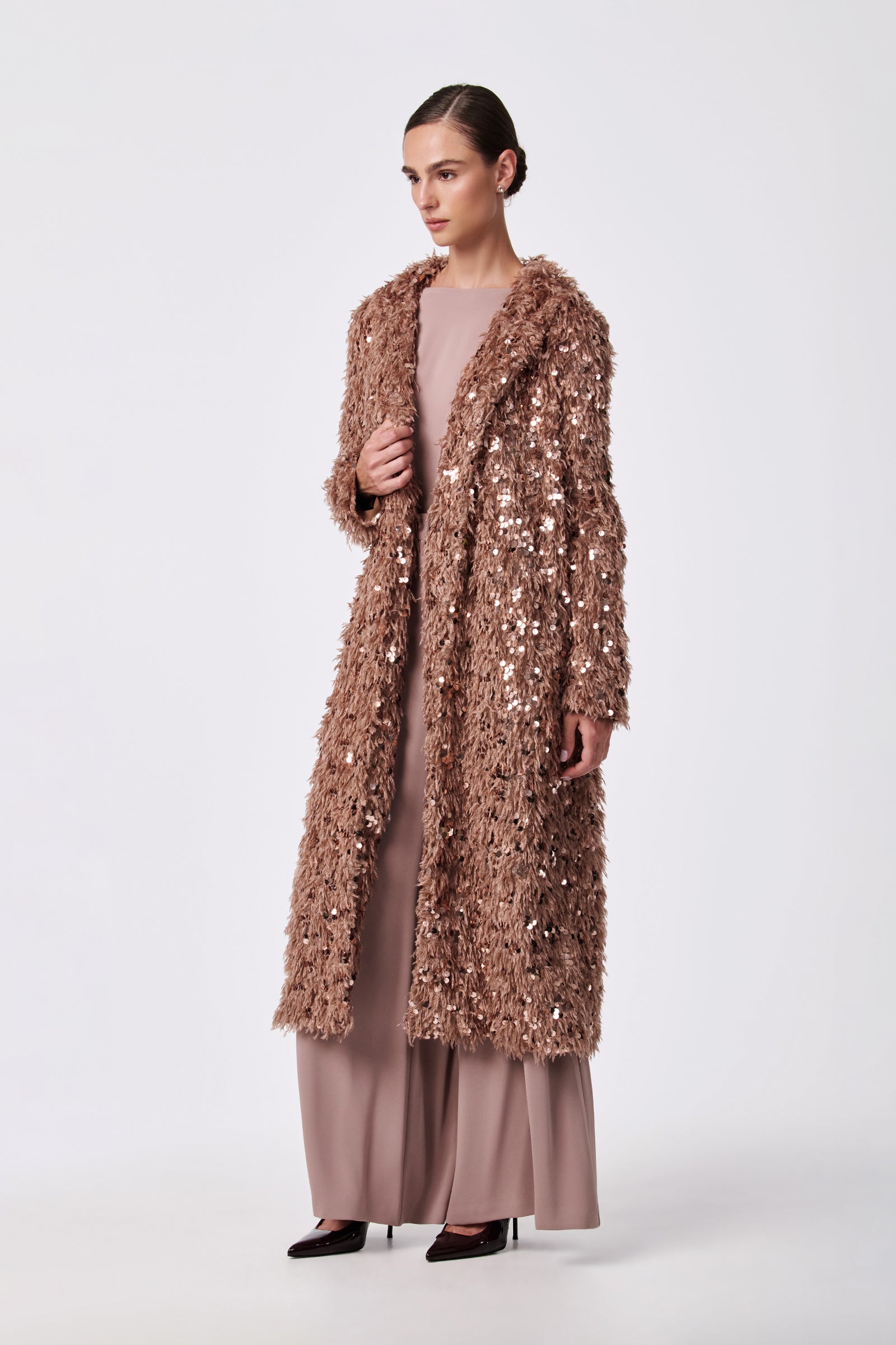 A woman stands against a plain background, wearing the Anna Coat—a long, textured rose gold sequin coat—over a mauve dress and dark pointed heels. Her hair is slicked back as she looks slightly to the side.