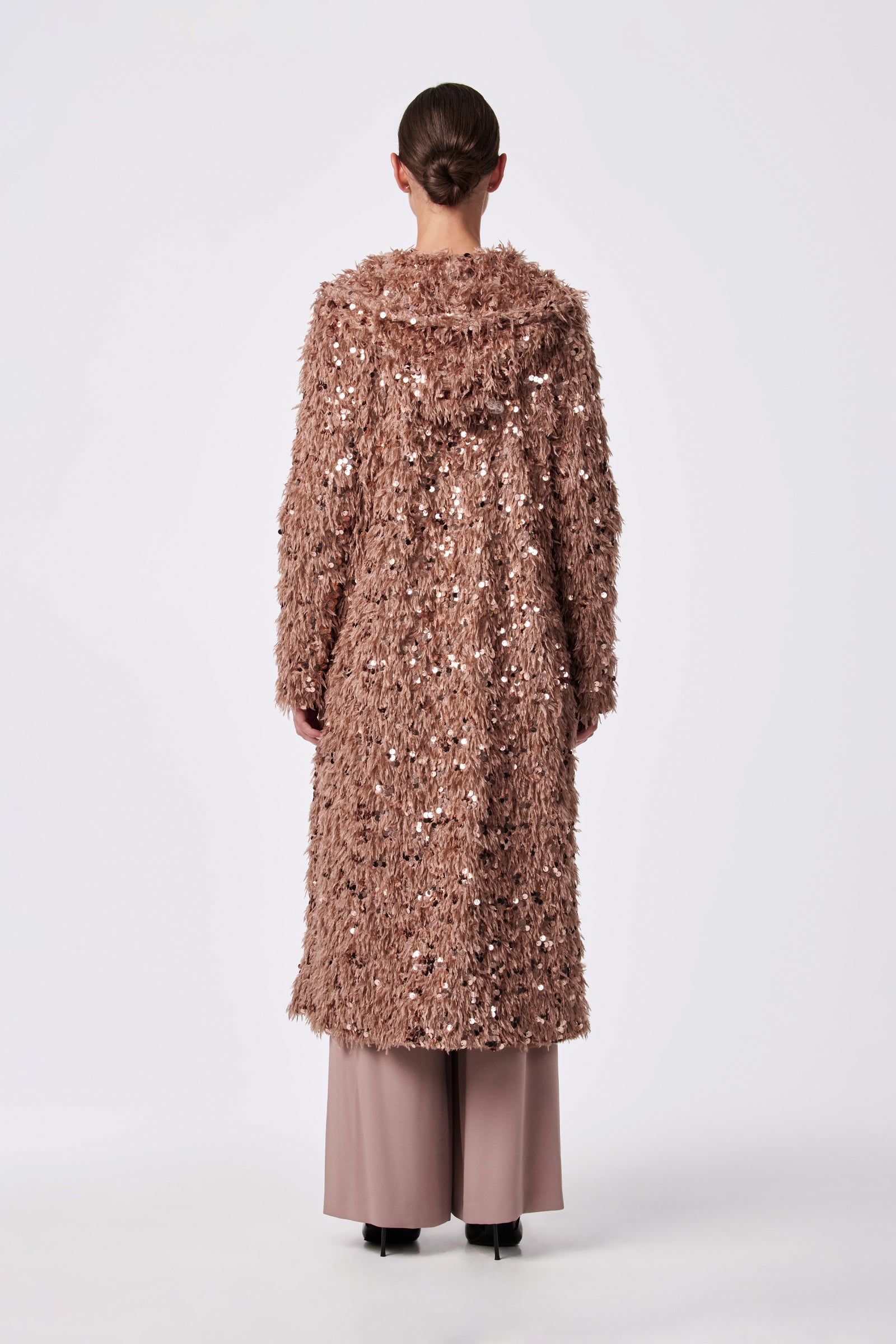 Model faces away, wearing the Anna Coat—a long, textured rose gold sequin layer—over wide-legged taupe pants and black shoes against a plain light background.