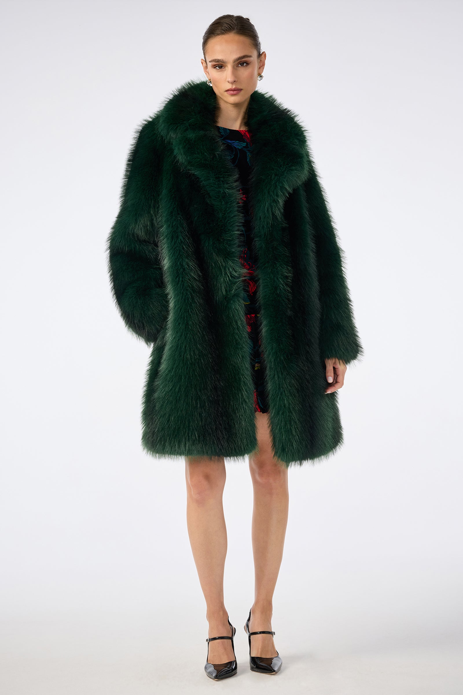 A woman stands against a plain background wearing the luxurious, oversized Dayos Coat in dark green faux fur, styled over a short dress with black heels. She gazes directly at the camera with a neutral expression.