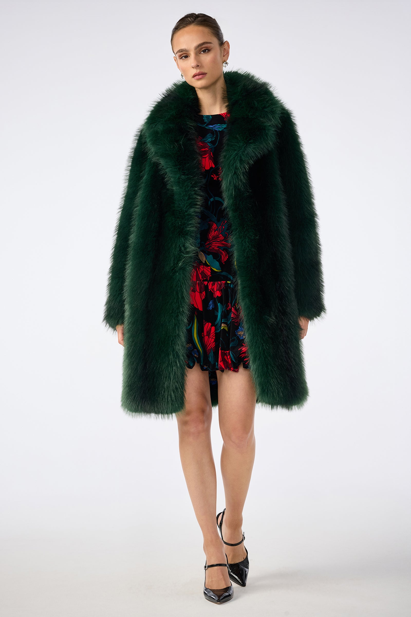 A woman stands against a plain white background wearing the Dayos Coat, a dark green faux fur piece, layered over a black dress with red floral patterns. Her hair is pulled back, and she completes the look with black pointed heels.