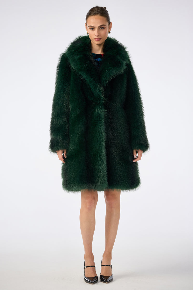 A woman faces forward in the Dayos Coat, a luxurious dark green faux fur design with a wide collar and long sleeves. She pairs it with black pointed-toe heels against a plain white background.