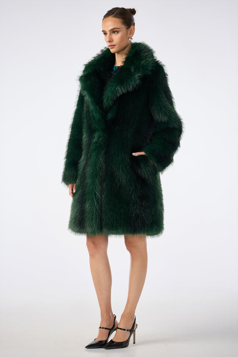 A woman with her hair in a bun stands against a plain background, wearing the Dayos Coat in dark green faux fur and black high heels. She has one hand in her pocket and a neutral expression.