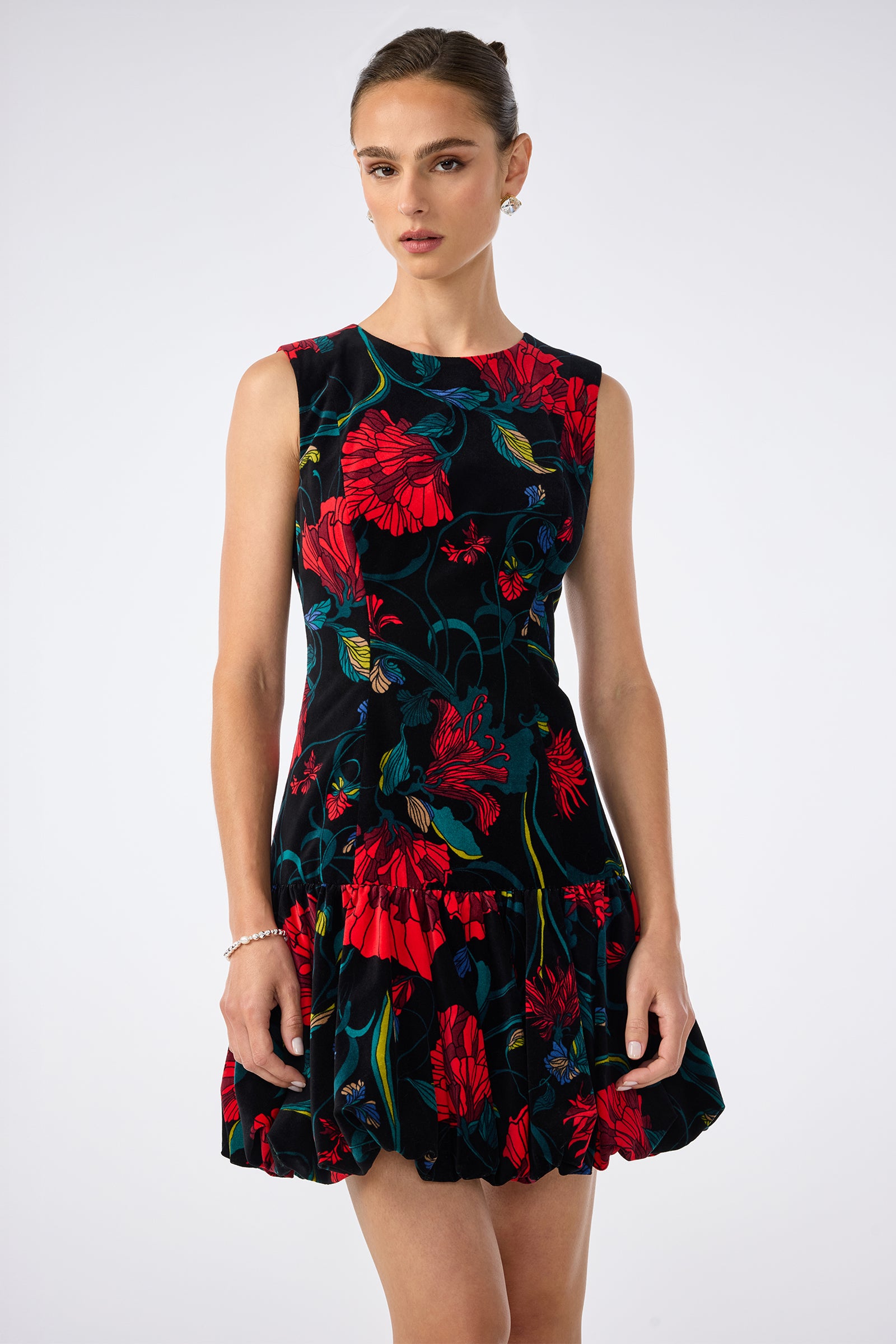 A woman wearing the Splendored Dress, a sleeveless black dress adorned with large red and blue floral patterns, stands against a plain light background.