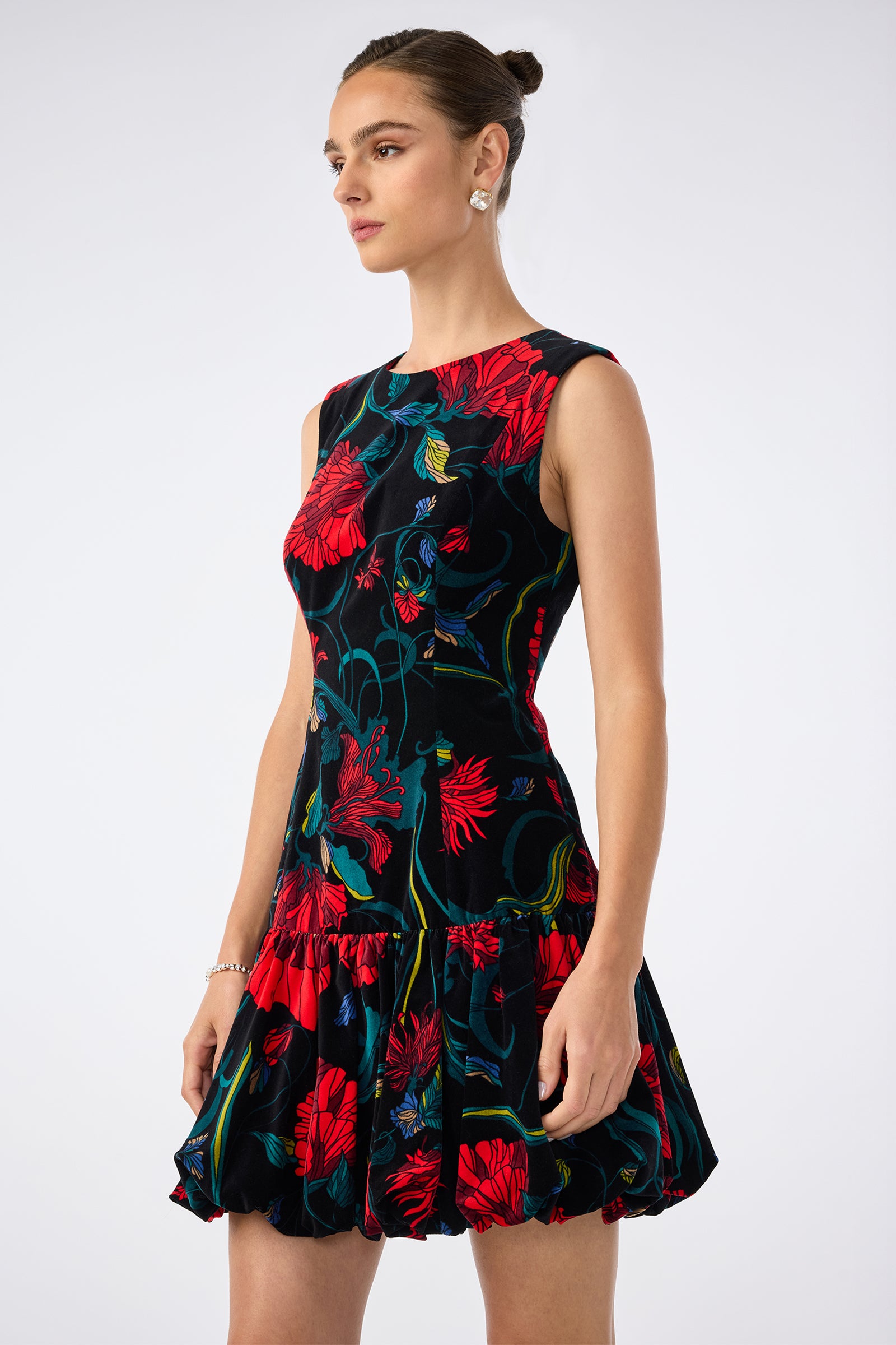 A woman models the Splendored Dress, a sleeveless black dress with red and blue floral print, fitted bodice, and flared ruffled hem, standing against a white background with her hair in a neat bun and wearing stud earrings.