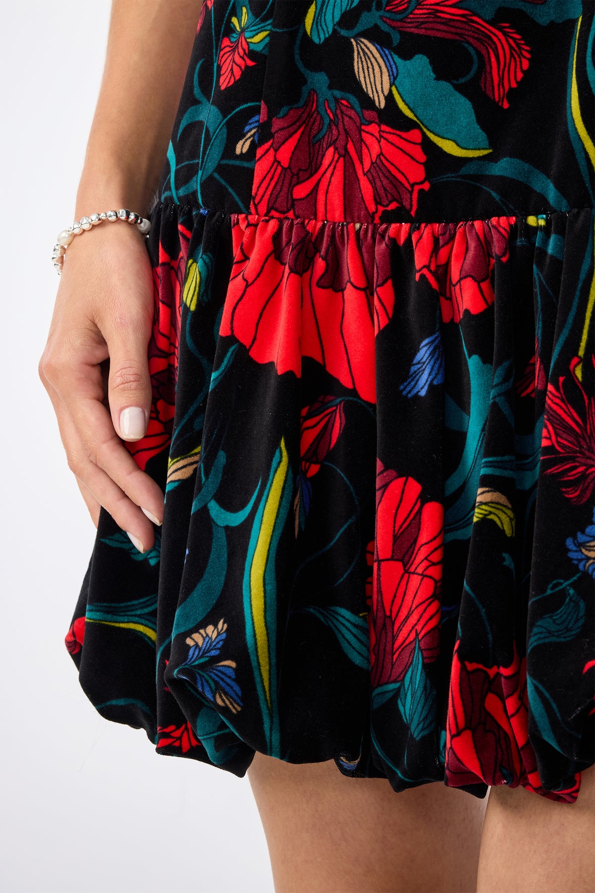 A close-up of a person in the Splendored Dress—a black dress with bold red, green, and blue floral designs. The individual has light skin, manicured nails, and is accessorized with a silver bracelet.