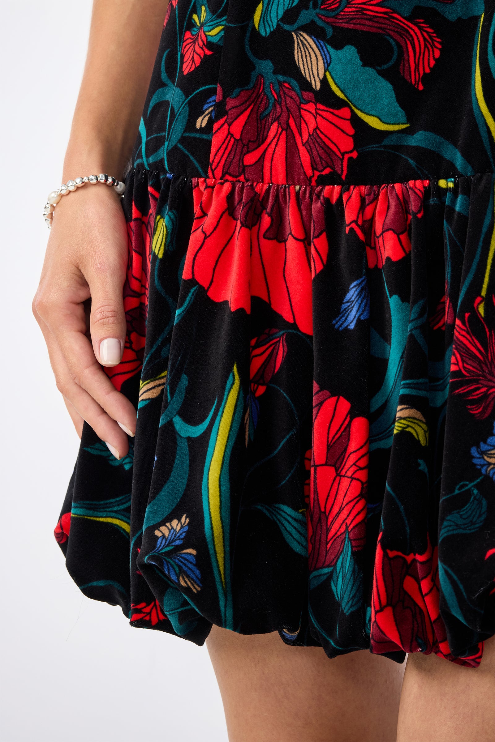 A close-up of a person in the Splendored Dress—a black dress with bold red, green, and blue floral designs. The individual has light skin, manicured nails, and is accessorized with a silver bracelet.