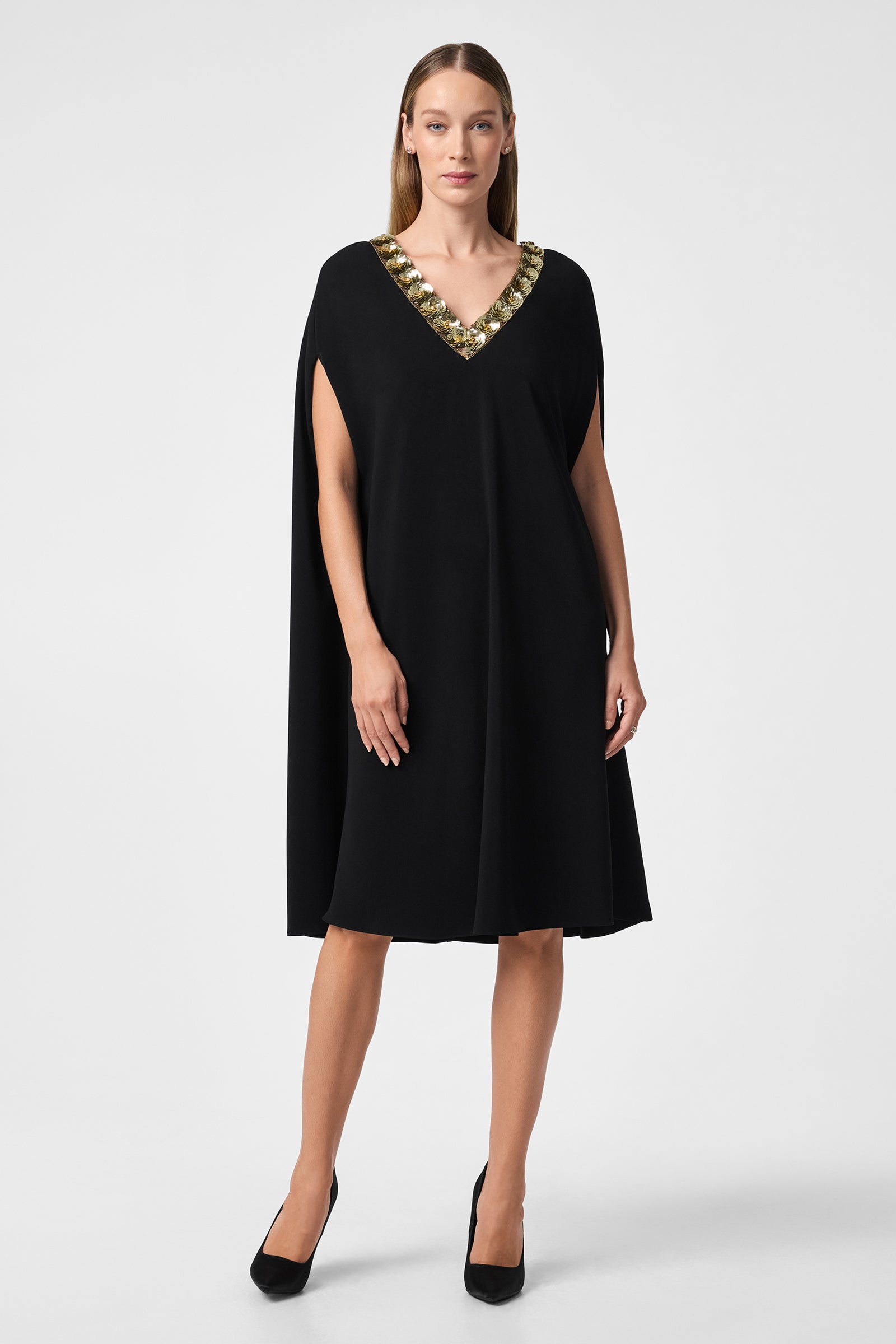 A woman stands facing forward in the Masquerade 3 Dress, a loose, knee-length black dress with a V-neckline featuring gold and silver embellishments. She has straight light brown hair, wears black heels, and poses against a white background.
