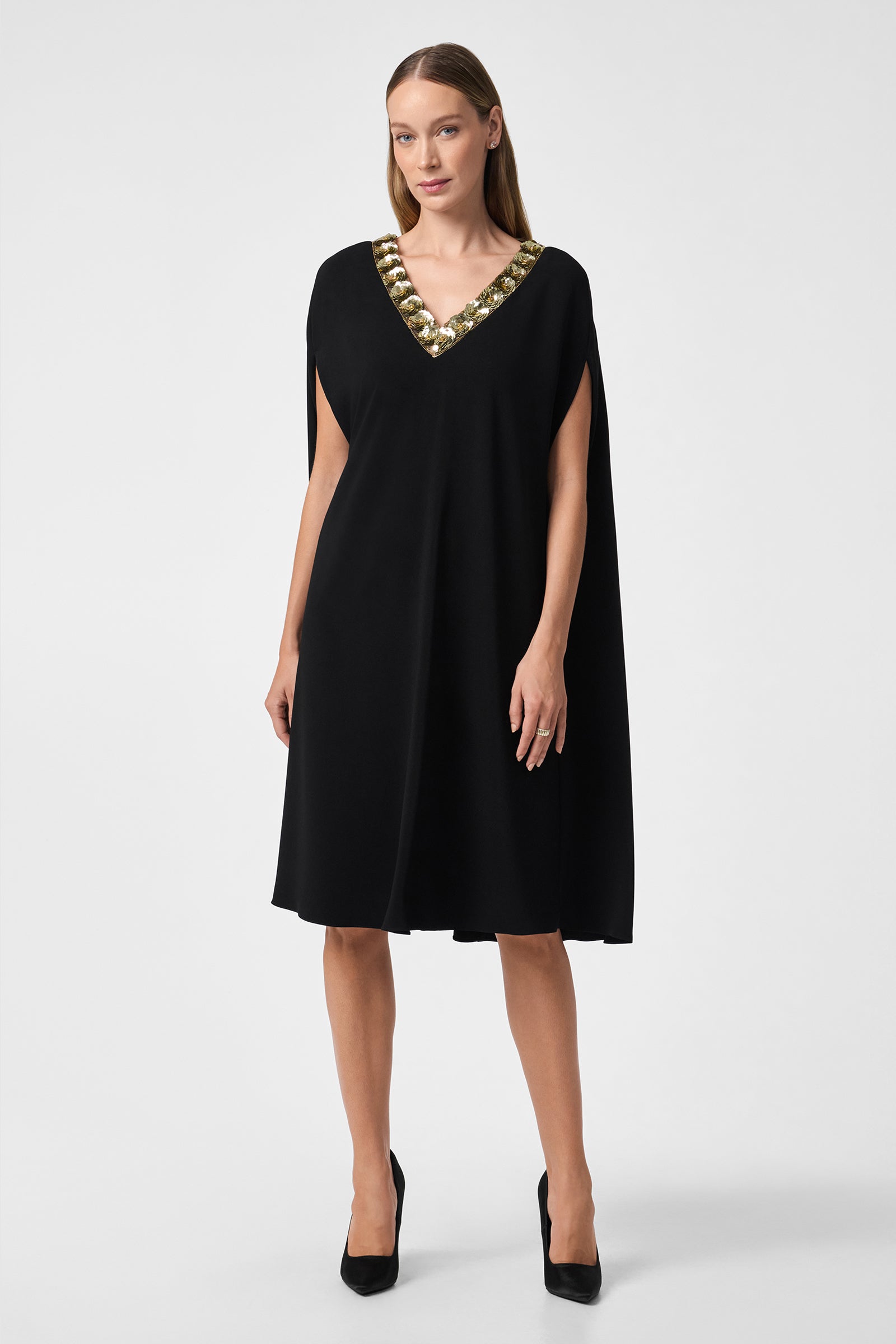 A woman stands against a plain white background wearing the Masquerade 3 Dress, a knee-length black dress with a gold embellished V-neckline, paired with black high heels.
