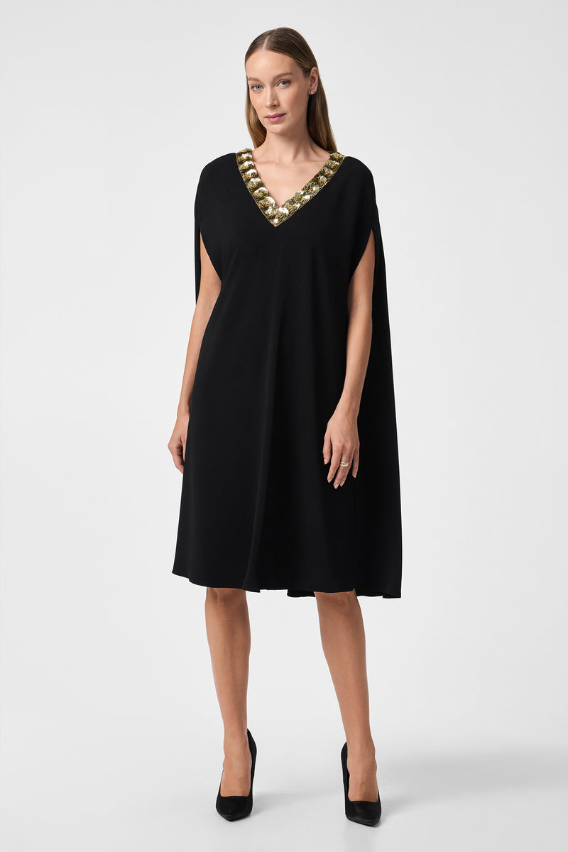 A woman stands against a plain white background wearing the Masquerade 3 Dress, a knee-length black dress with a gold embellished V-neckline, paired with black high heels.