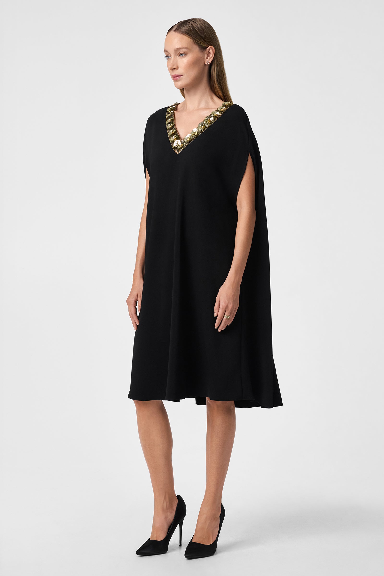A woman wears the Masquerade 3 Dress, a knee-length, loose-fitting black dress with a gold-embellished V-neckline. She has straight light brown hair, black heels, and stands against a plain light background.