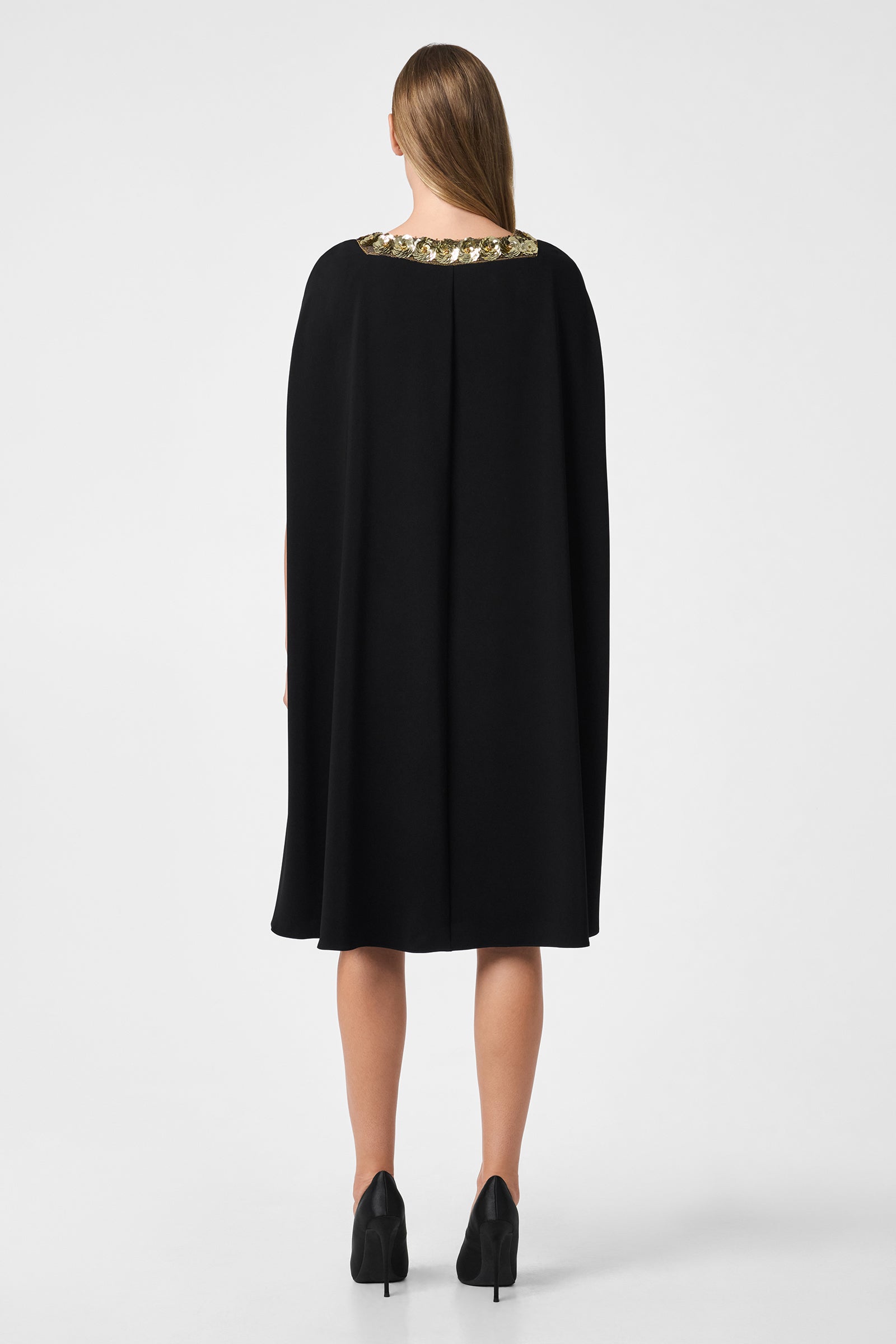 A woman faces away, wearing the Masquerade 3 Dress—a knee-length black cape dress with an embellished neckline—and black heels. Her straight hair is down against a plain white background.