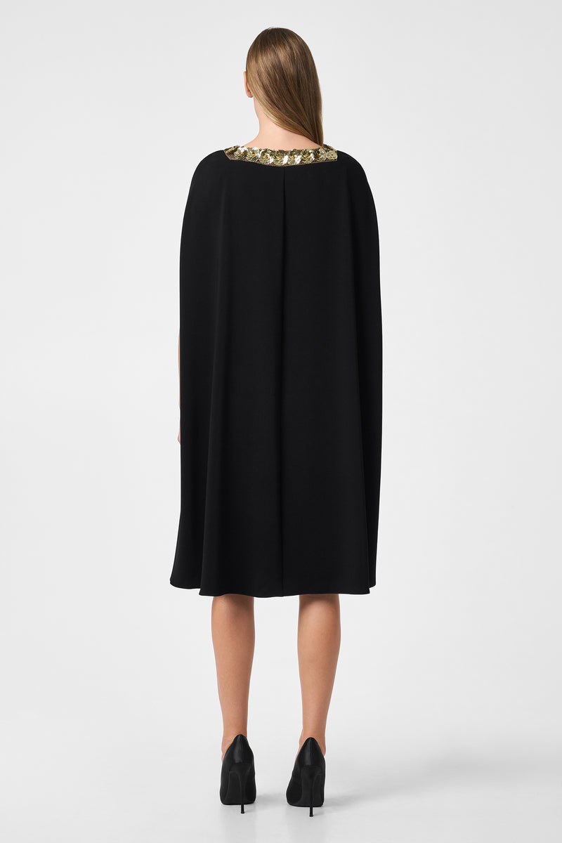 A woman faces away, wearing the Masquerade 3 Dress—a knee-length black cape dress with an embellished neckline—and black heels. Her straight hair is down against a plain white background.