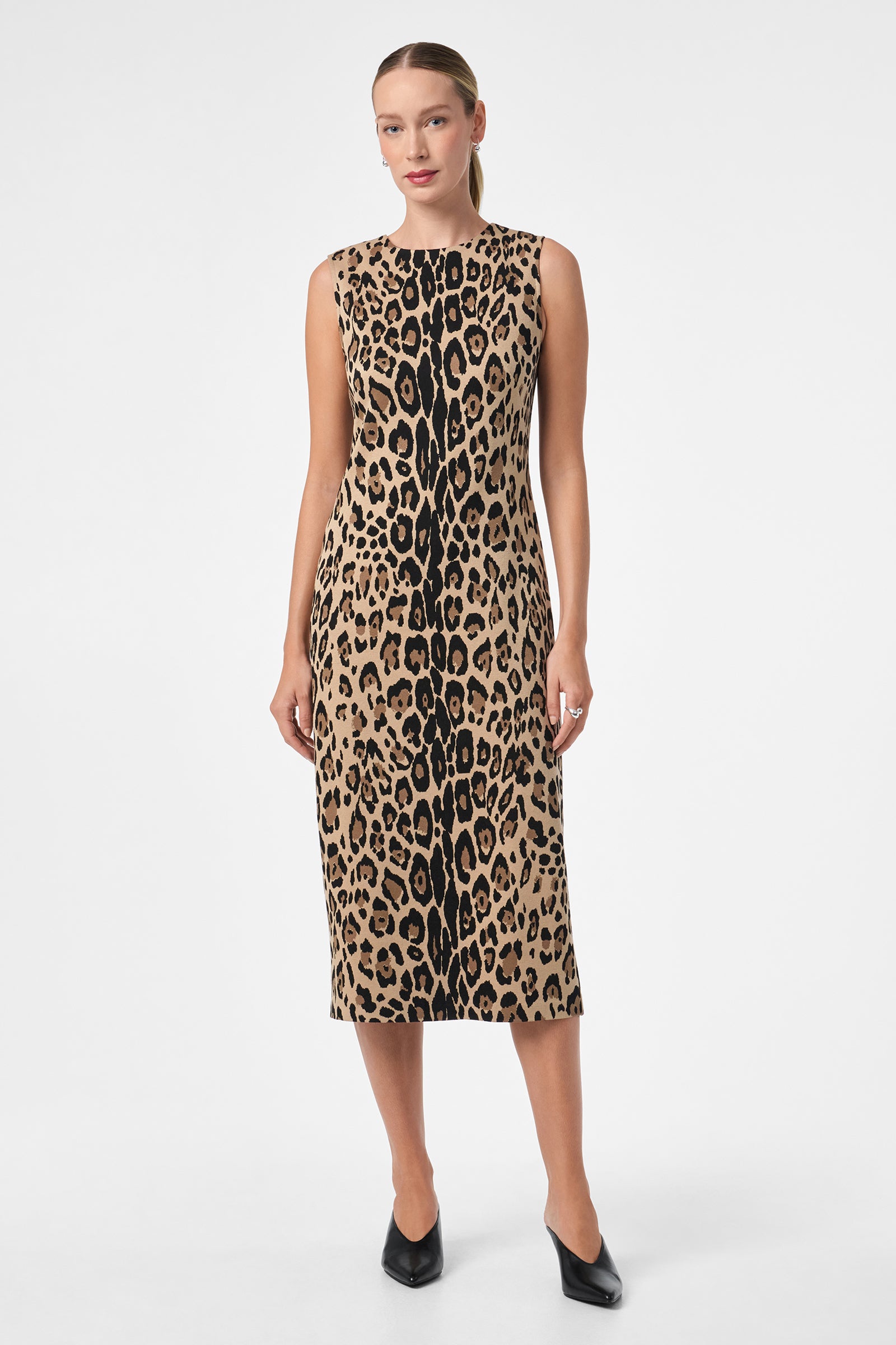 A woman poses against a white background wearing the Dame Dress, a sleeveless, knee-length, form-fitting piece featuring a bold brown and black leopard print. She completes the look with black heels.