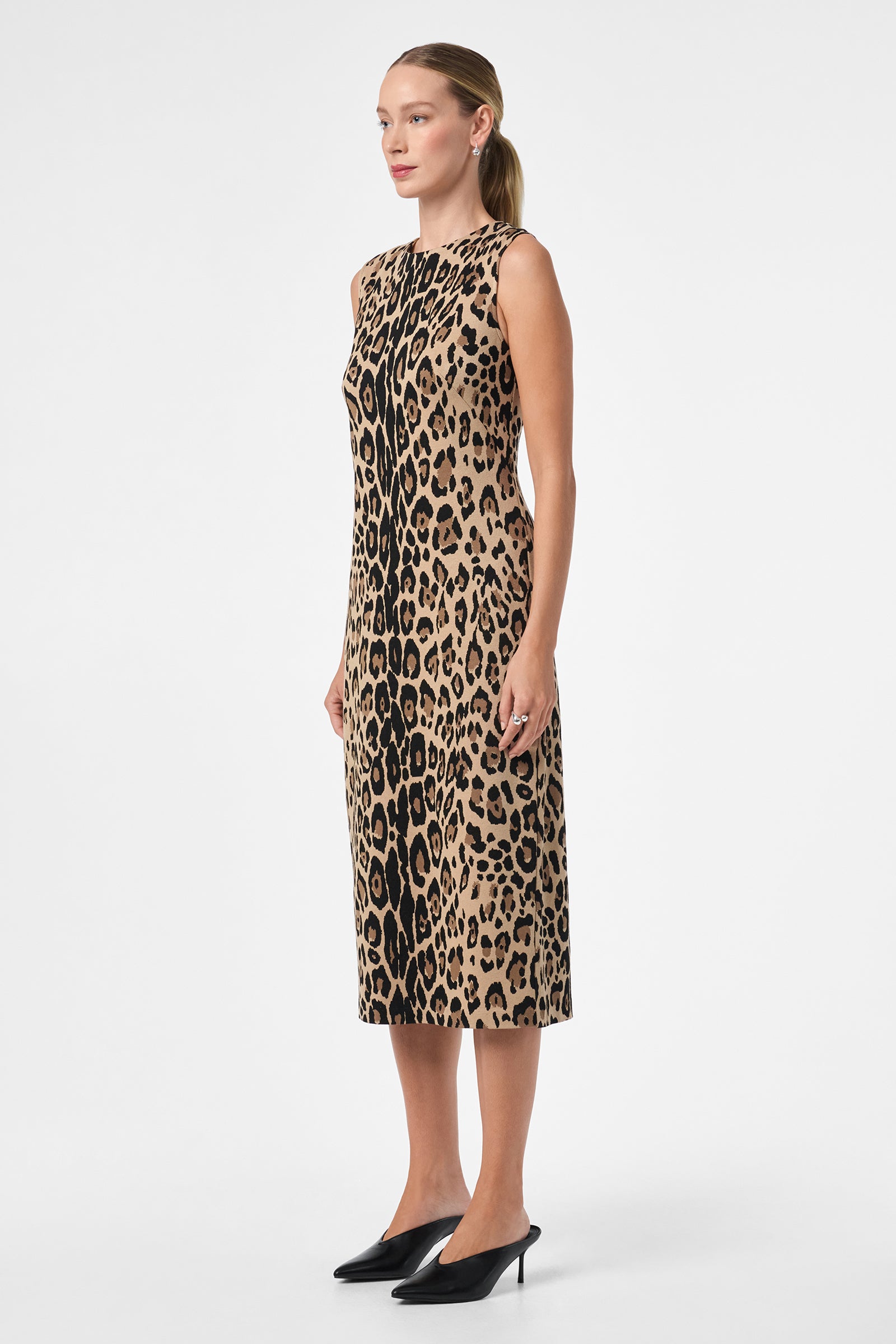 A woman with straight, pulled-back hair stands against a plain white background wearing the sleeveless, knee-length Dame Dress in leopard print and black heels, facing slightly to the side.