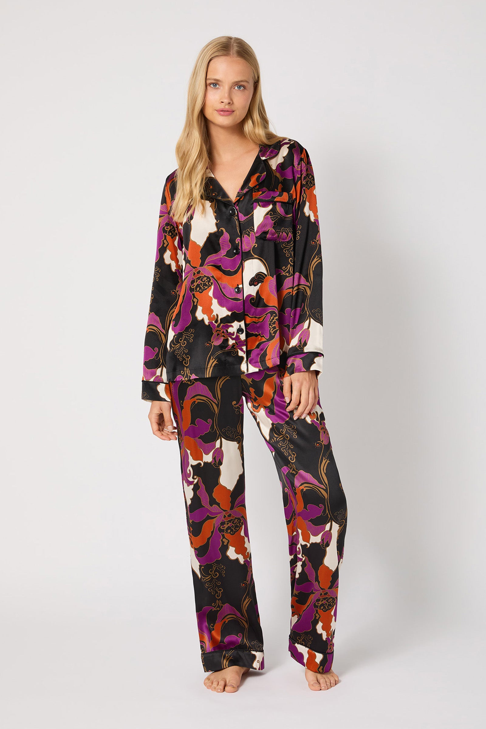 A woman with long blonde hair stands barefoot against a plain background, wearing the Classic Silk PJ Set - Fall Floral, featuring a bold, colorful abstract pattern in black, purple, orange, pink, and white.