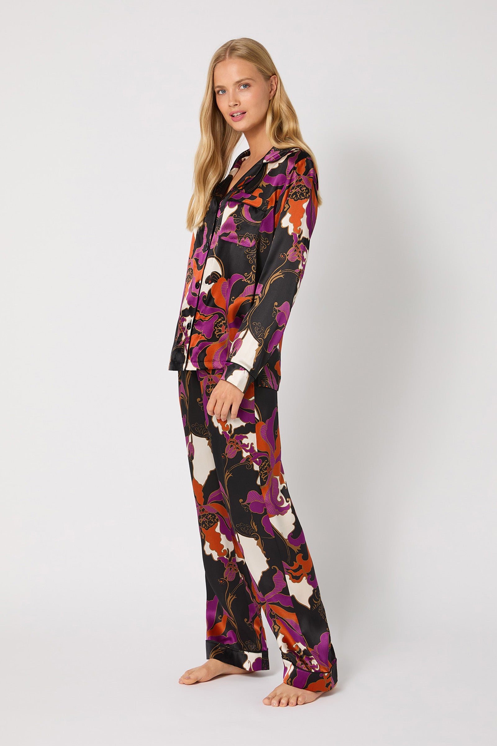 A woman with long blonde hair stands barefoot against a plain backdrop, wearing the Classic Silk PJ Set - Fall Floral featuring a bold abstract pattern in black, purple, orange, and white. She looks at the camera with a slight smile.