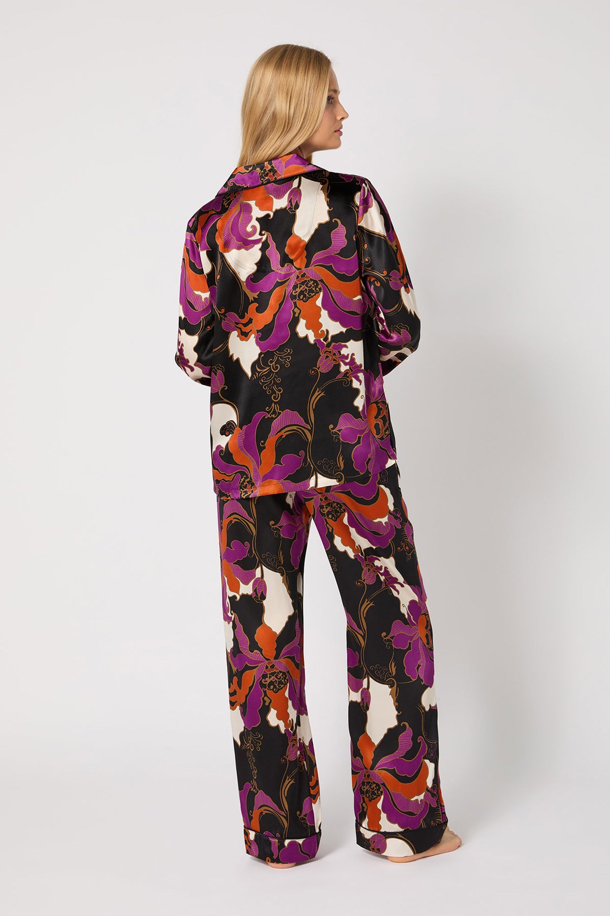 A woman stands barefoot facing away, wearing the Classic Silk PJ Set - Fall Floral: loose-fitting matching satin pajamas with a bold purple, orange, white, and black floral print. The background is plain white.