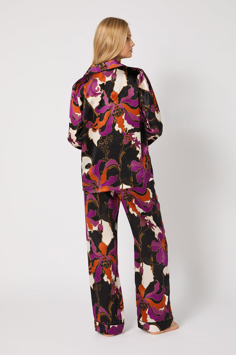 A woman stands barefoot facing away, wearing the Classic Silk PJ Set - Fall Floral: loose-fitting matching satin pajamas with a bold purple, orange, white, and black floral print. The background is plain white.