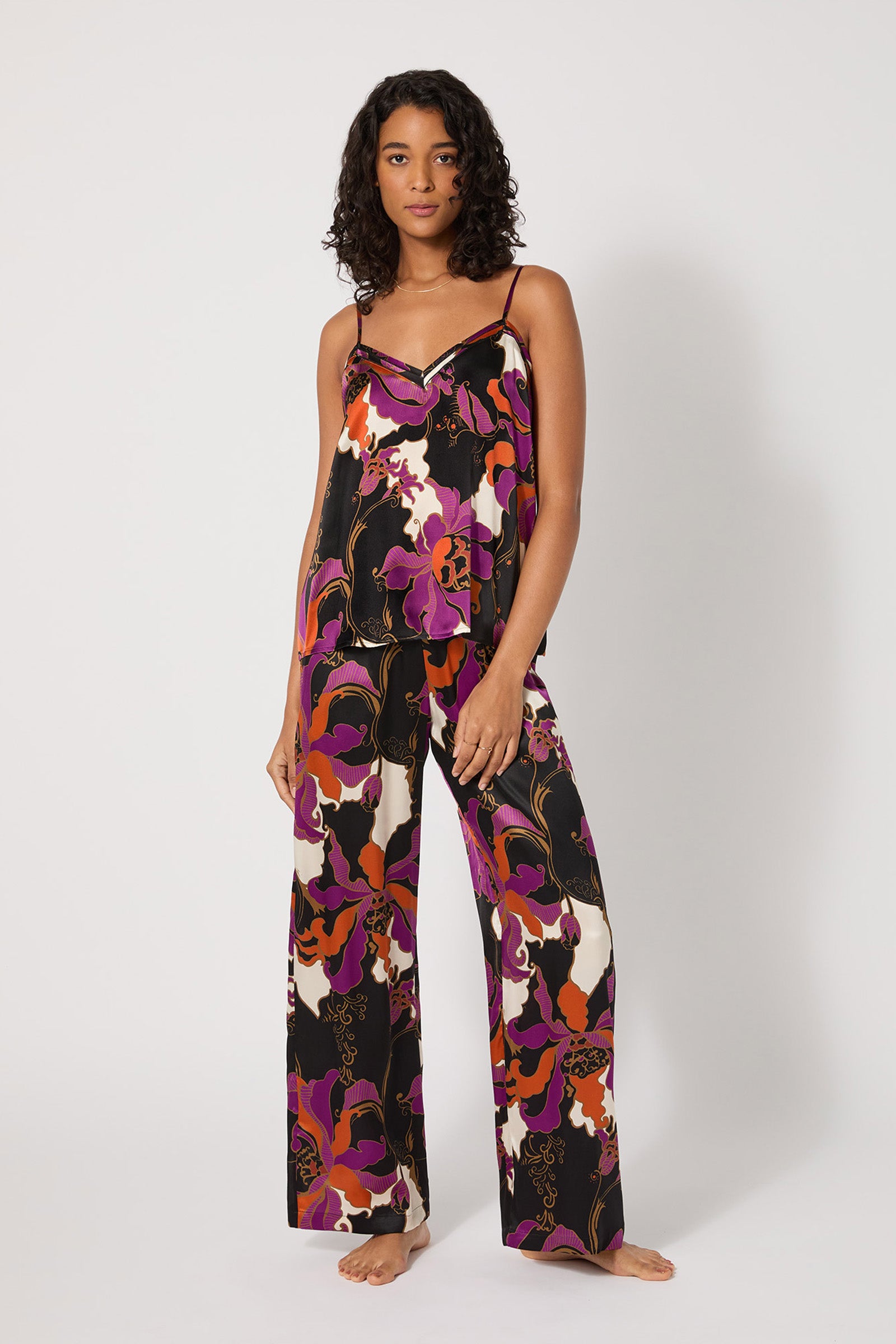 A woman stands barefoot against a white background, wearing the Silk Cami PJ Set - Fall Floral, featuring a black base with bold pink, purple, red, and orange floral patterns. The set includes a sleeveless top and wide-leg pants.