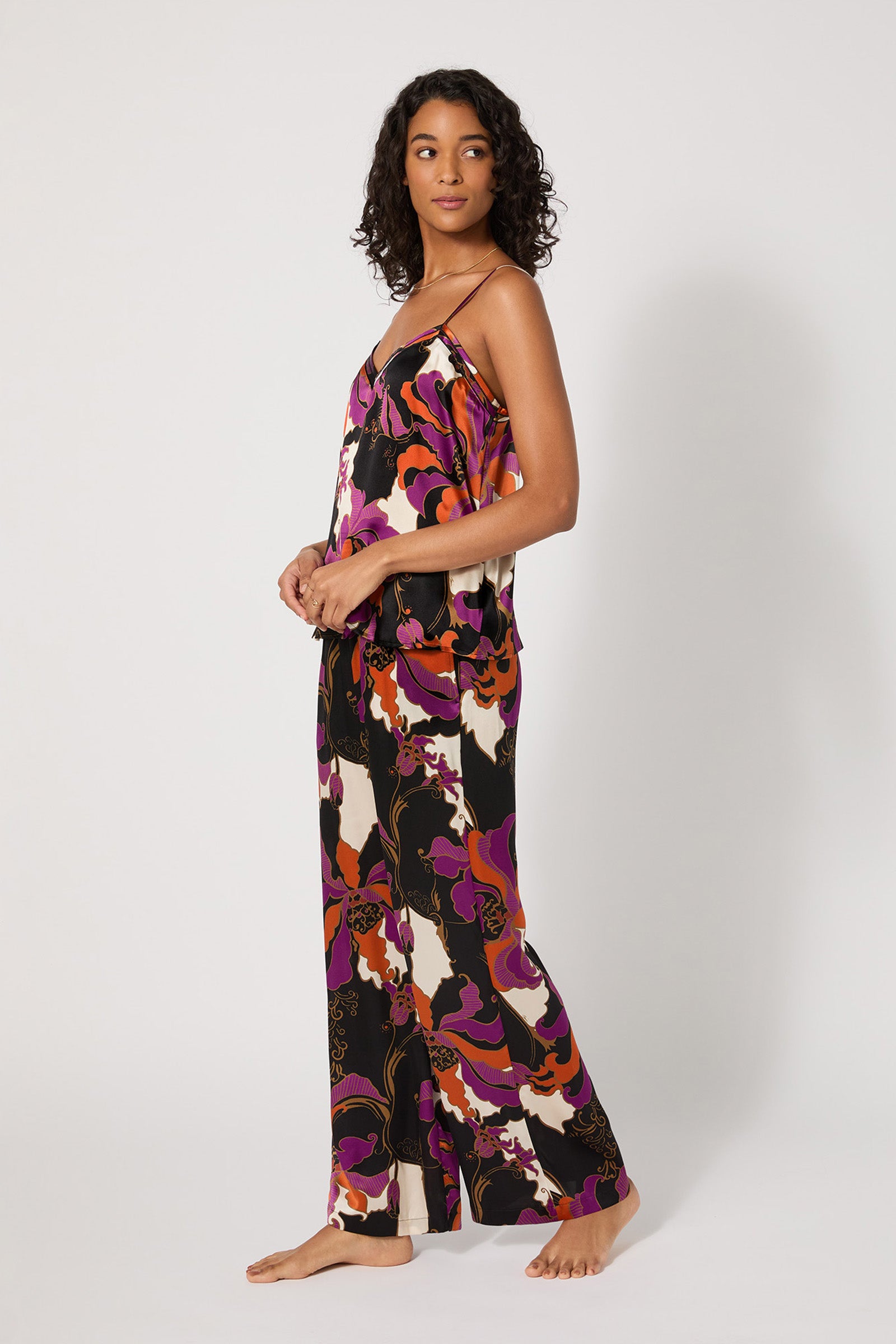 A woman stands barefoot against a plain background, wearing the Silk Cami PJ Set - Fall Floral, a loose matching pajama set with a bold abstract floral pattern in purple, orange, black, and white. She gazes to the side with a relaxed expression.
