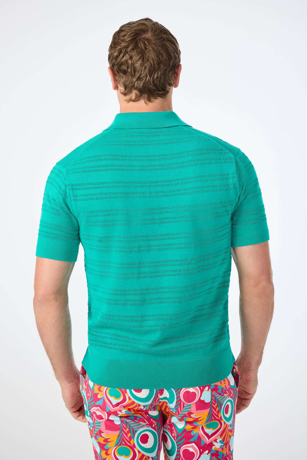 A person with short, light brown hair is seen from behind, wearing the Edison Short-Sleeve Striped Polo and colorful abstract-patterned pants featuring red, yellow, white, and teal shapes on a white background.
