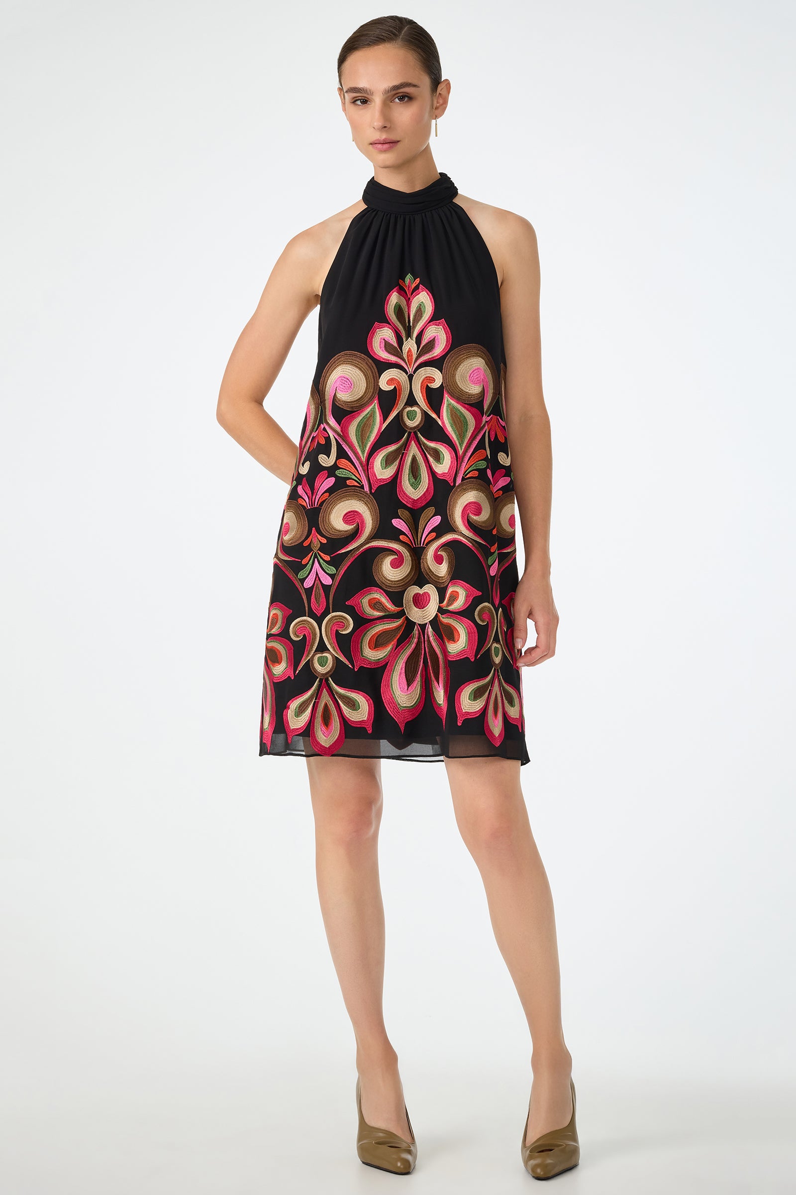 A woman stands against a plain white background wearing the Zana Embroidered Mini Dress in Sanctuary, a sleeveless, knee-length black dress with bold pink, red, and green floral embroidery, paired with bronze heels.