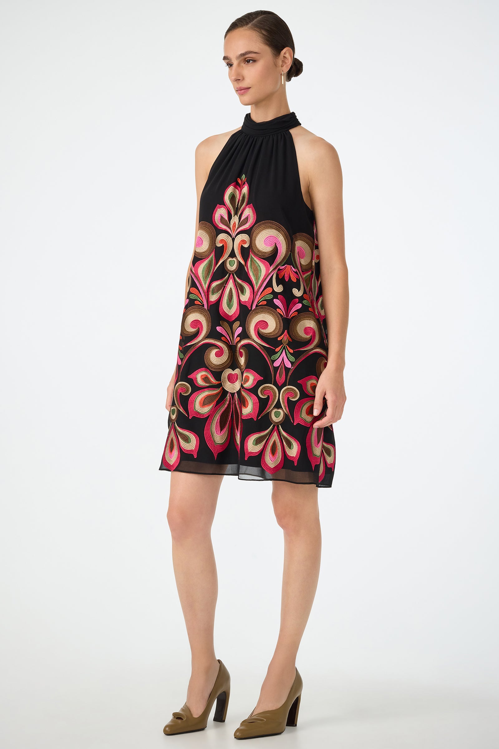 A woman wears the Zana Embroidered Mini Dress, featuring a black halter-neck design with ornate pink, red, and gold floral embroidery, paired with tan high heels against a plain white background.