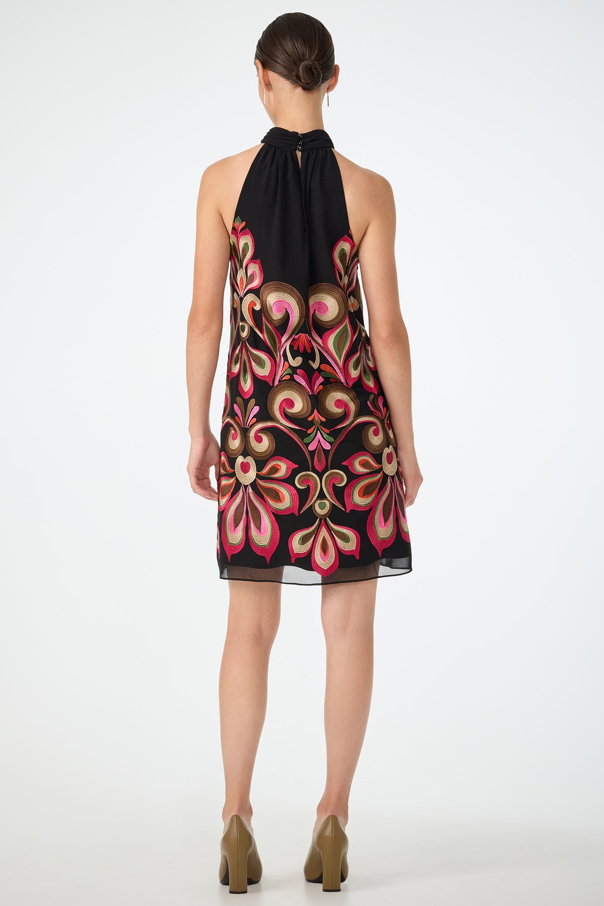 A woman faces away, wearing the Zana Embroidered Mini Dress—a sleeveless black dress with bold pink and gold floral embroidery. Her hair is tied back, and she pairs the look with beige block-heeled shoes against a plain white background.