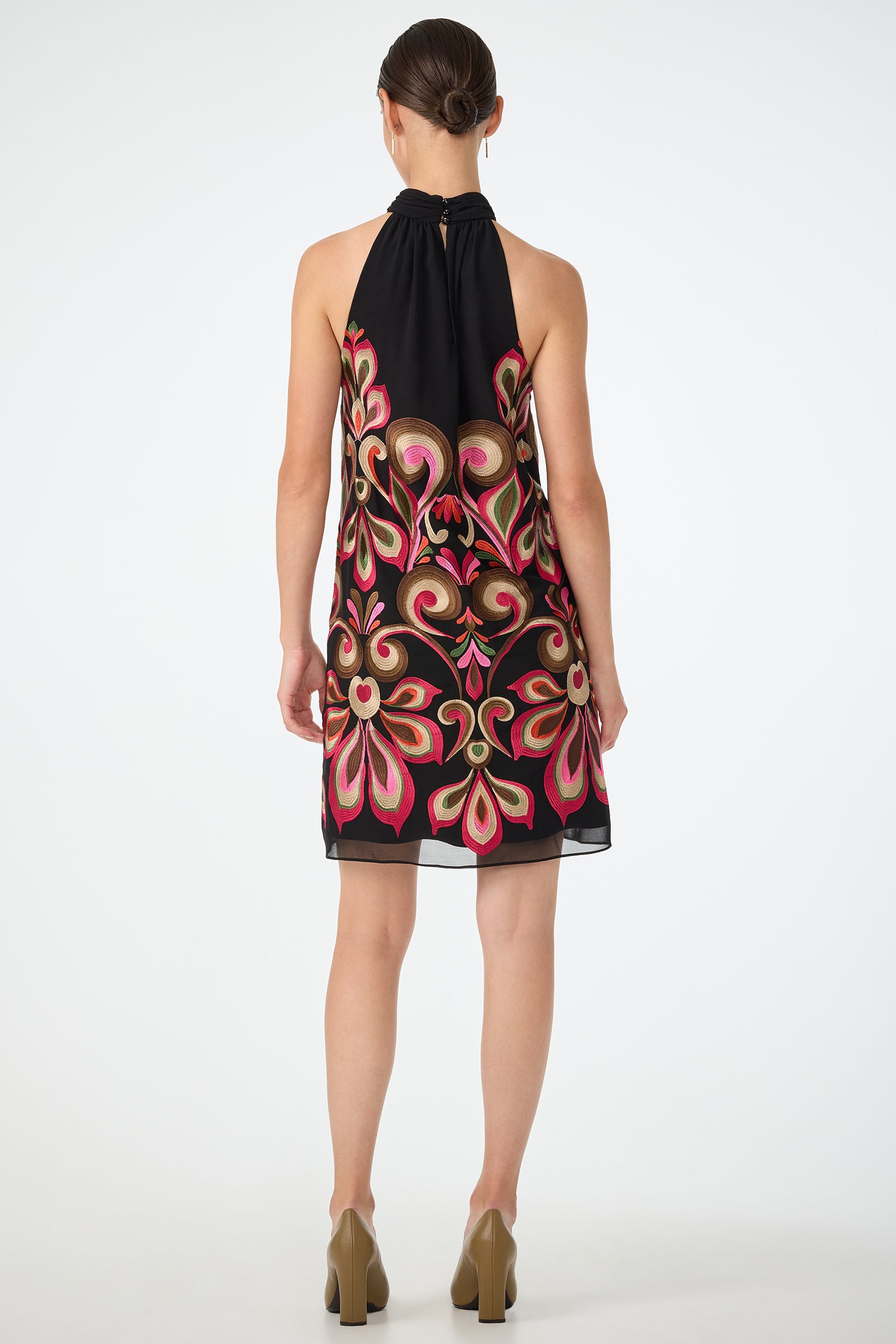 A woman faces away, wearing the Zana Embroidered Mini Dress—a sleeveless black dress with bold pink and gold floral embroidery. Her hair is tied back, and she pairs the look with beige block-heeled shoes against a plain white background.