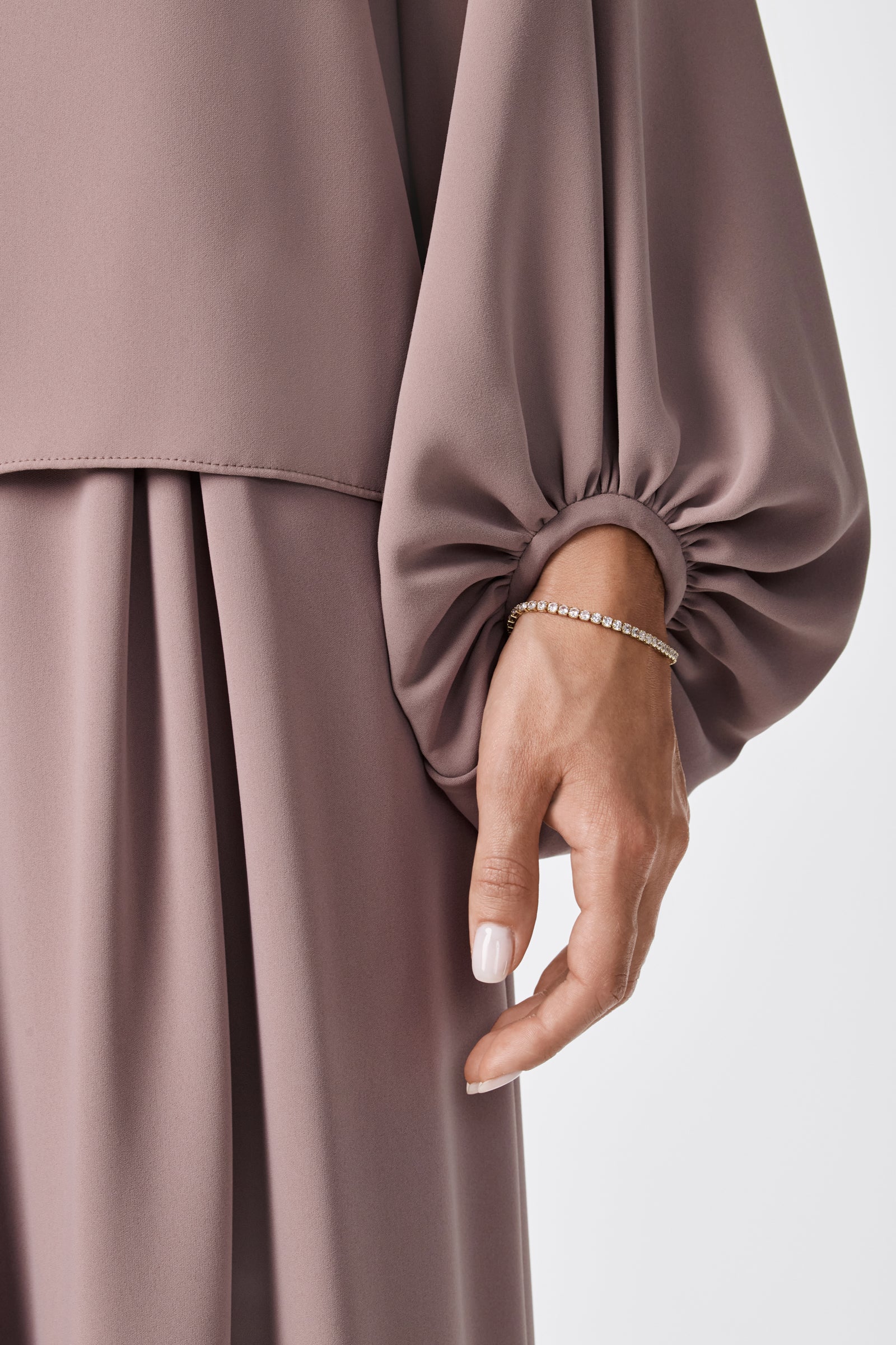 A close-up of a person wearing the URSA 2 TOP—a mauve, long-sleeve blouse with gathered cuffs made from soft polyester—paired with a matching skirt. Light-colored nails and a thin beaded bracelet complete the elegant look.