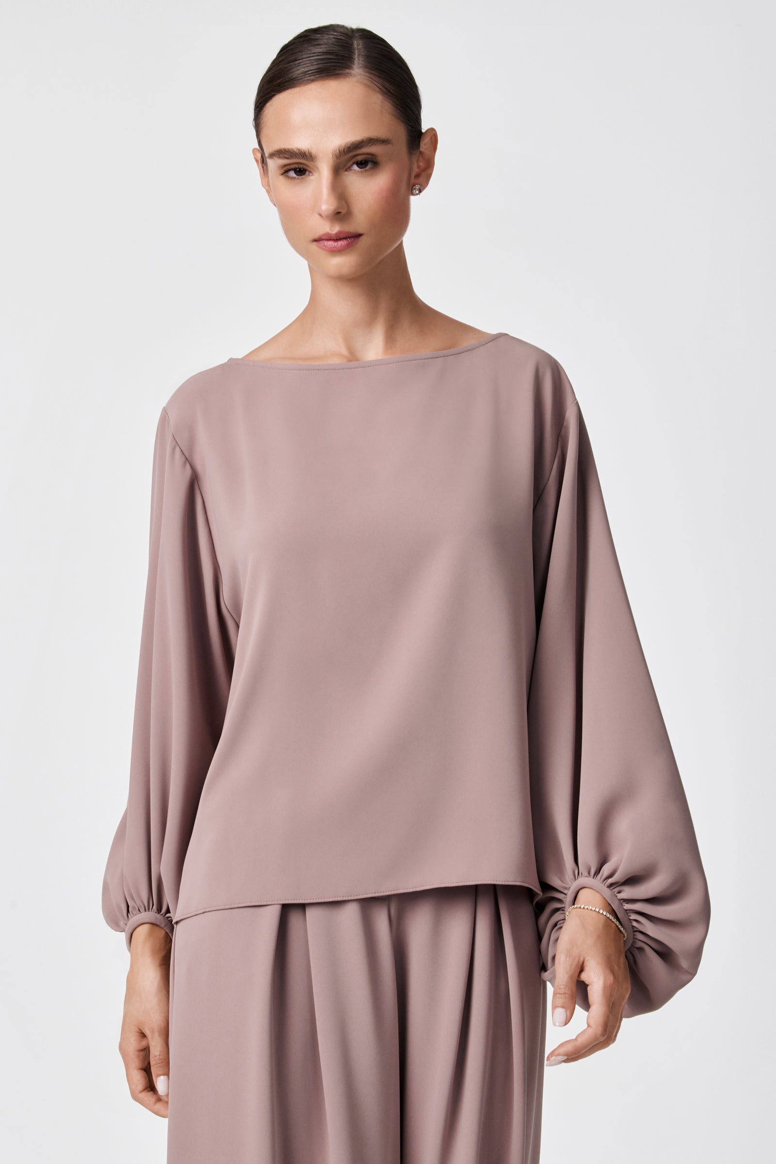 A woman stands against a plain light background wearing the URSA 2 TOP, a loose-fitting, long-sleeved mauve blouse with gathered cuffs, paired with matching wide-leg pants, all crafted from soft polyester fabric.