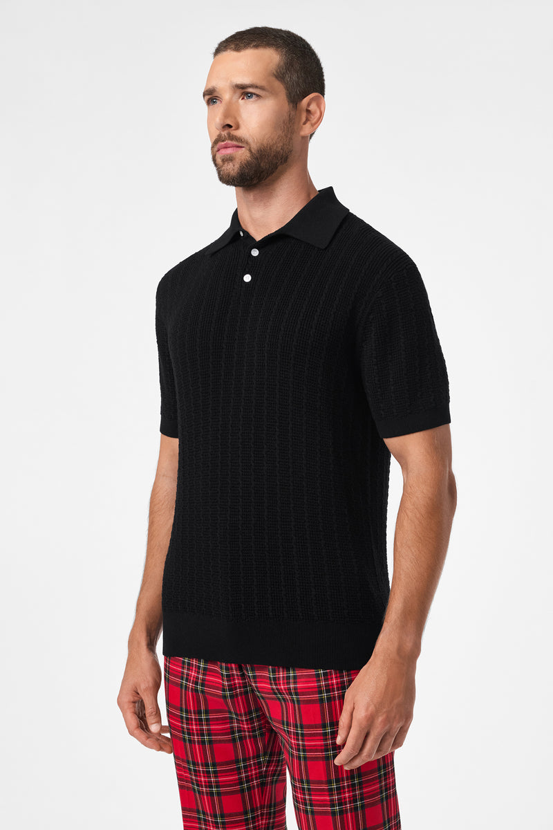 A man with short hair and a beard wears the Asher Short-Sleeve Polo in black and red plaid pants, standing against a plain white background.