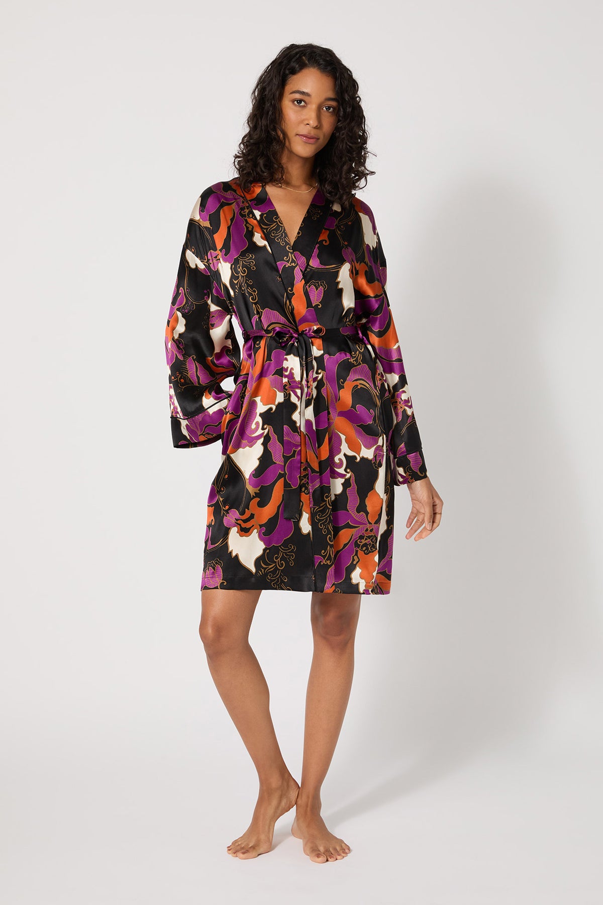 A woman stands barefoot against a plain background, wearing the Long-Sleeve Silk Robe - Fall Floral in multi, a knee-length silk robe with a vibrant floral pattern, long sleeves, and a waist tie.