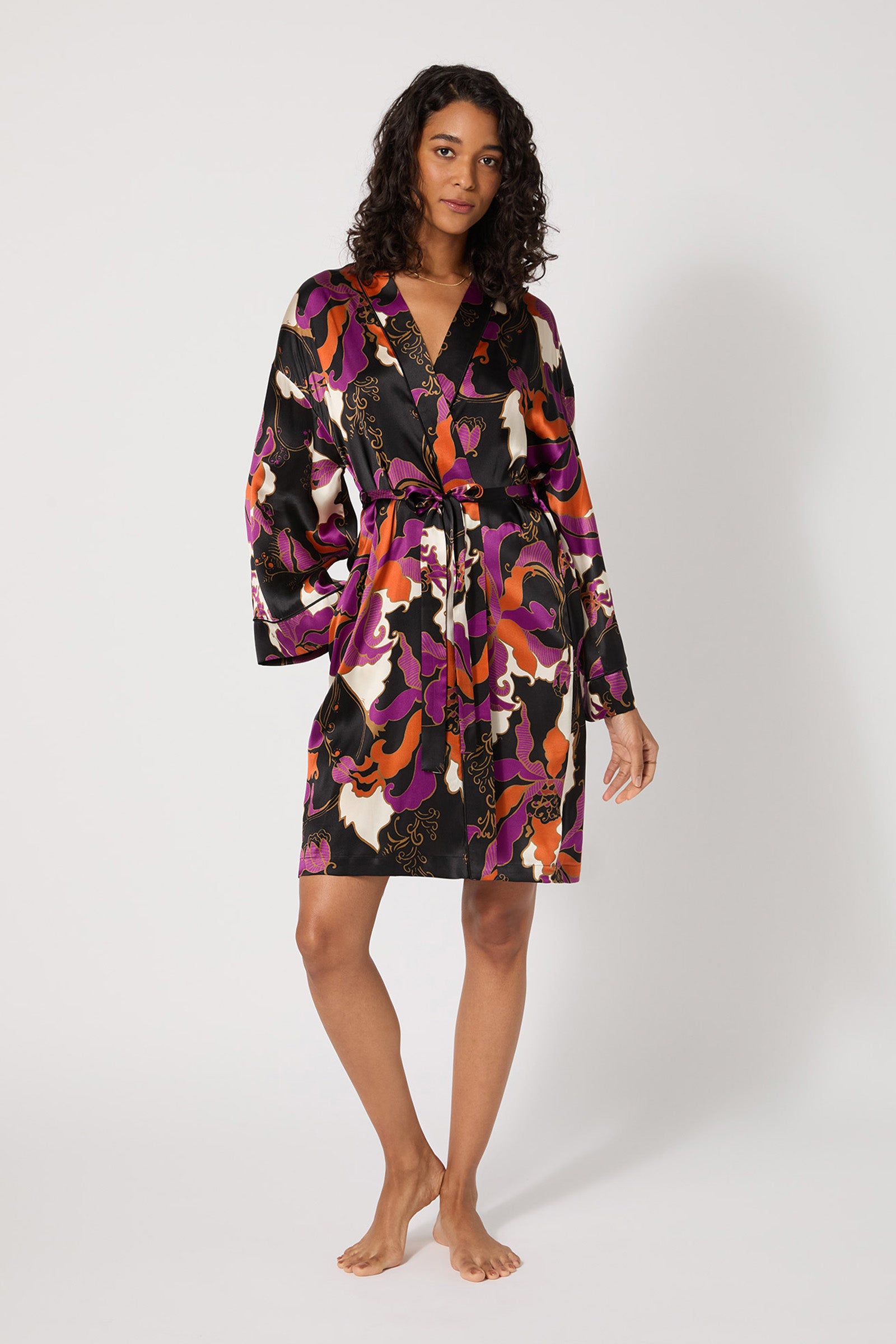 A woman stands barefoot against a plain background, wearing the Long-Sleeve Silk Robe - Fall Floral in multi, a knee-length silk robe with a vibrant floral pattern, long sleeves, and a waist tie.