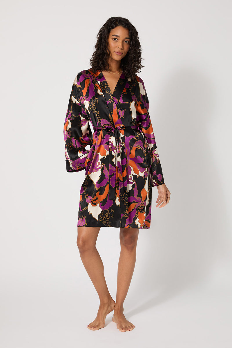 A woman stands barefoot against a plain background, wearing the Long-Sleeve Silk Robe - Fall Floral in multi, a knee-length silk robe with a vibrant floral pattern, long sleeves, and a waist tie.