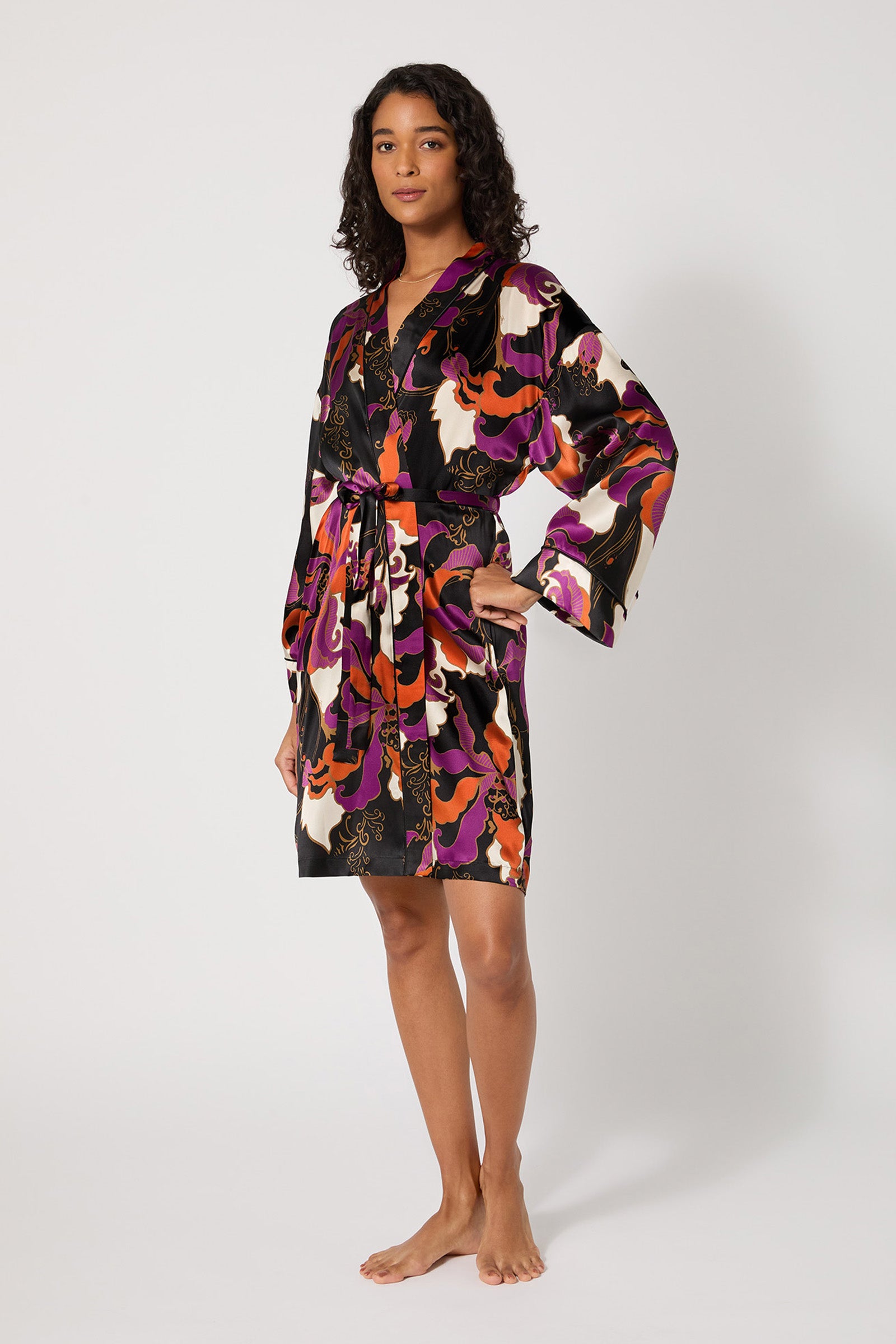 A woman stands barefoot against a white background, wearing the Long-Sleeve Silk Robe - Fall Floral, featuring a bold purple, orange, pink, and black abstract pattern with a matching tie at the waist.