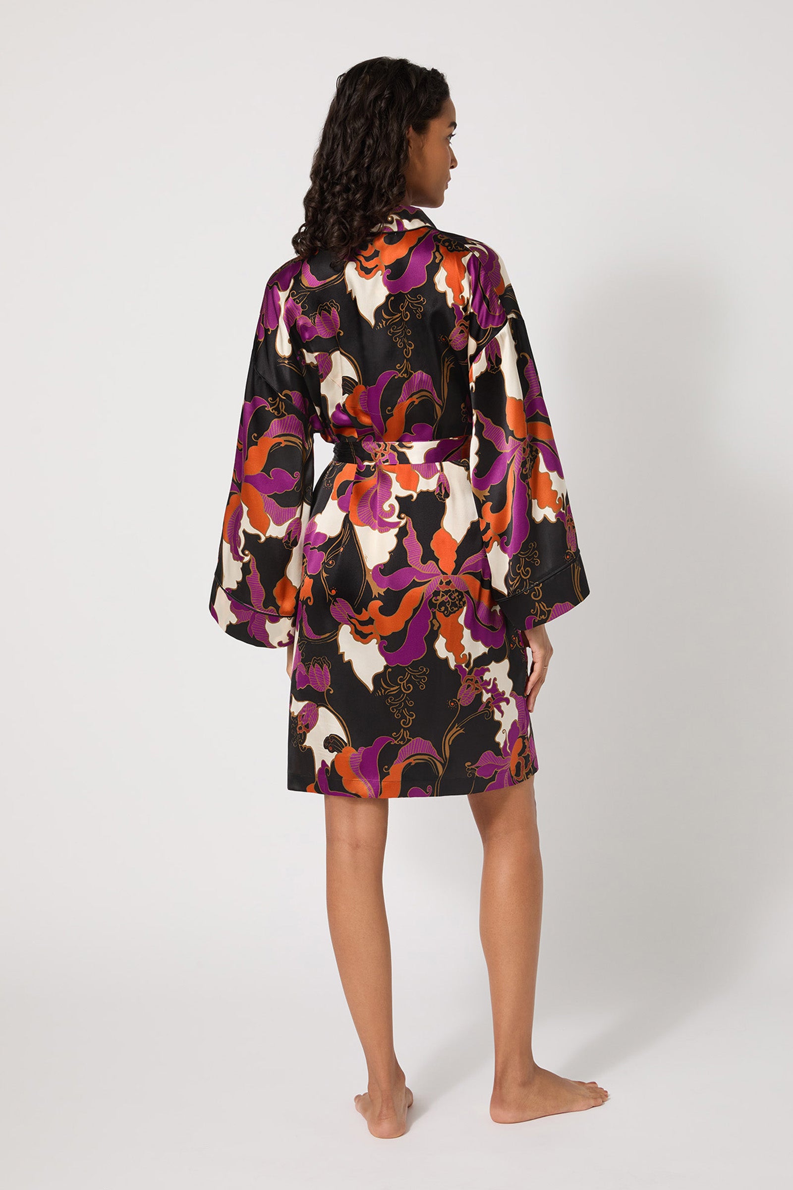 A woman with curly hair stands barefoot, facing away, wearing the Long-Sleeve Silk Robe - Fall Floral featuring a bold purple, black, white, and orange floral pattern against a plain white background.