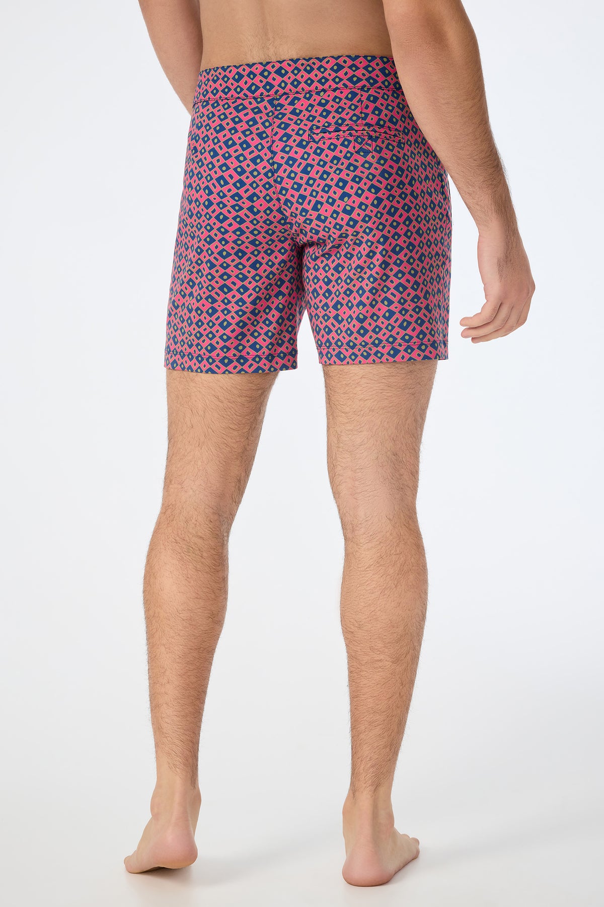 A man stands barefoot on a white background, shown from the back, wearing the Honolulu Board Short - Himari Herringbone with a colorful red, blue, and beige geometric pattern.