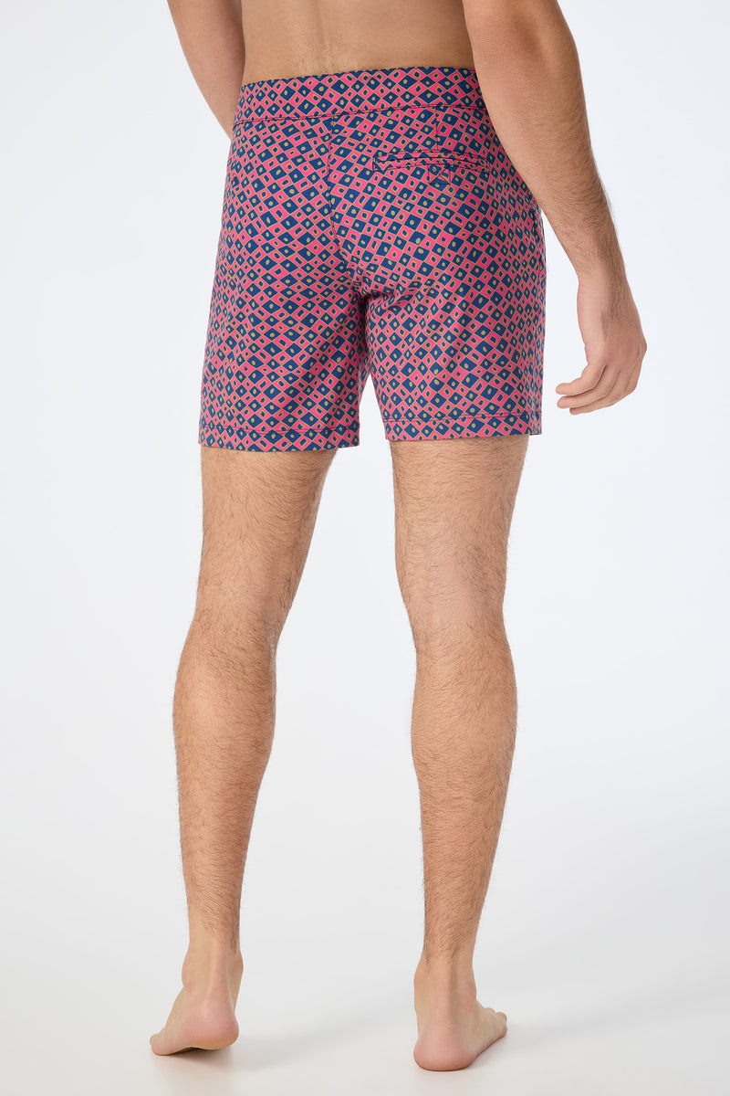 A man stands barefoot on a white background, shown from the back, wearing the Honolulu Board Short - Himari Herringbone with a colorful red, blue, and beige geometric pattern.