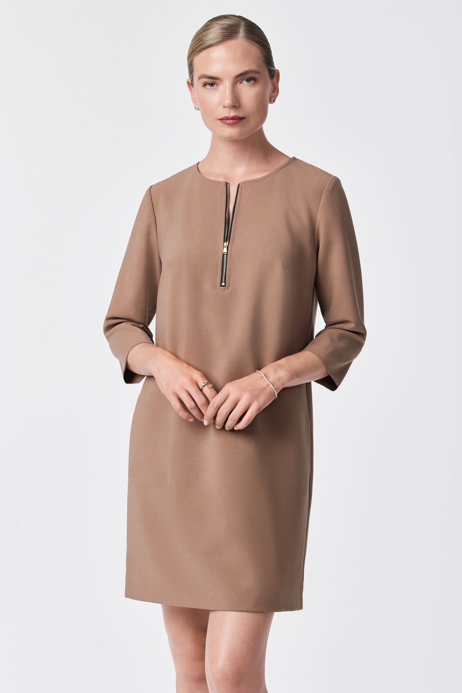 A woman with blonde hair in a neat bun wears the Versed Dress—a simple, knee-length tan dress with three-quarter sleeves and a front zipper—standing against a plain white background.