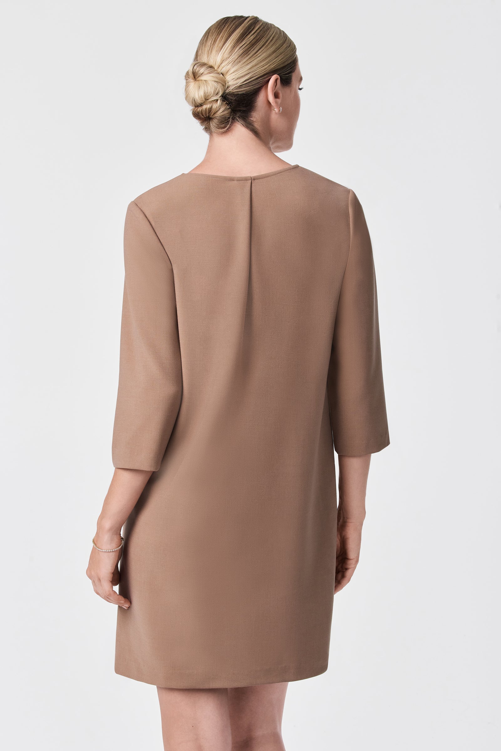 A woman with blonde hair in a neat low bun models the Versed Dress, a knee-length, camel-colored piece with three-quarter sleeves, shown from the back against a plain white background.