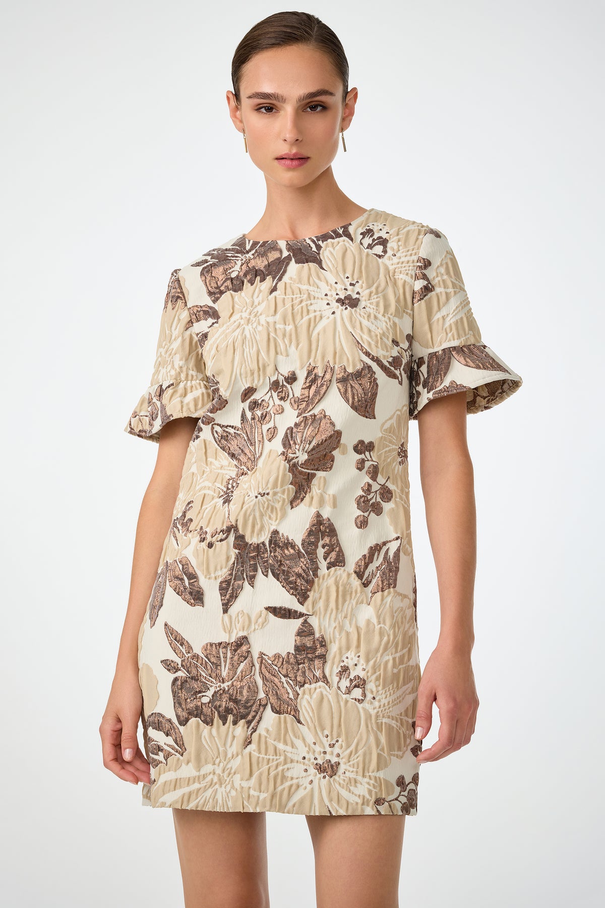 A woman wears the Darling Flounce Sleeve Dress, a short cream dress with bold brown and beige floral prints, flutter sleeves, and a round neckline, while standing against a plain white background.